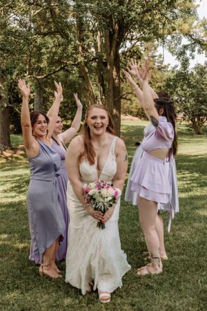 Bridesmaids in purple dresses celebrate with raised arms in sunny garden setting.