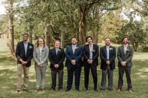 Wedding party members in suits stand in line against natural outdoor backdrop.