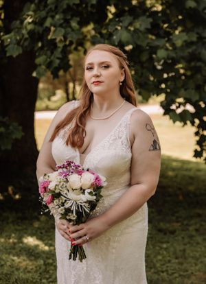 A person in a fitted lace wedding dress poses with a pink and white floral bouquet outdoors.