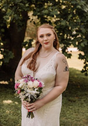A person in a lace wedding dress holds a pink and white bouquet while posing outdoors.