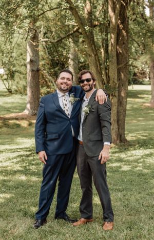 Wedding party members pose casually in a park with dappled sunlight filtering through trees.