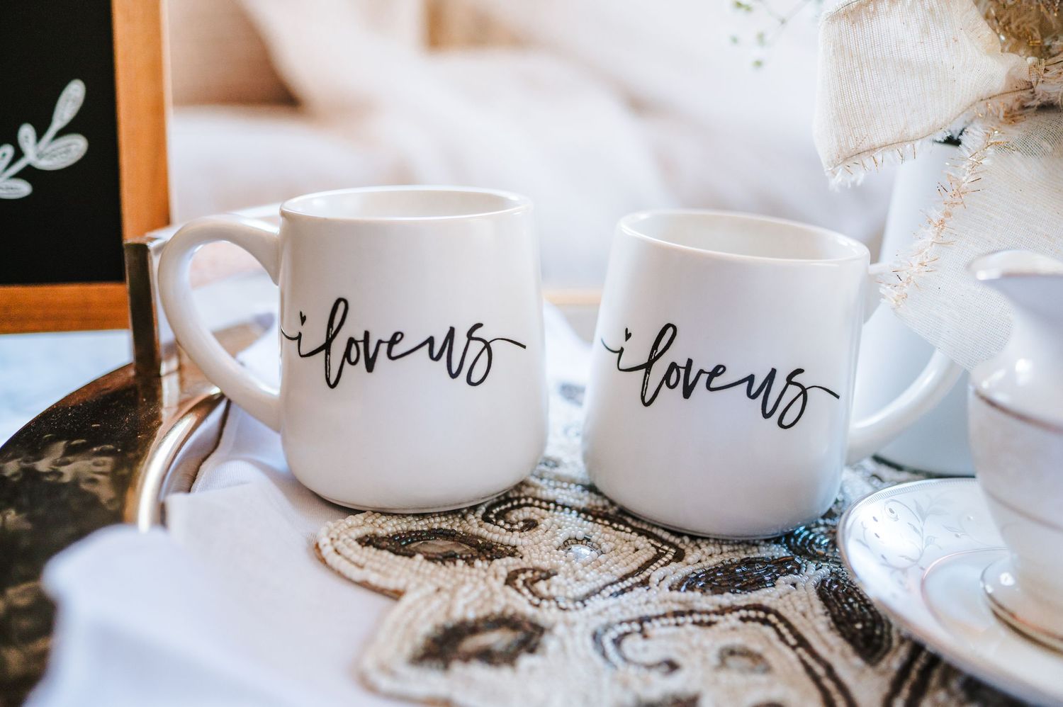 White ceramic coffee mugs with handwritten love text sit on a rustic wooden table with scattered coffee beans.