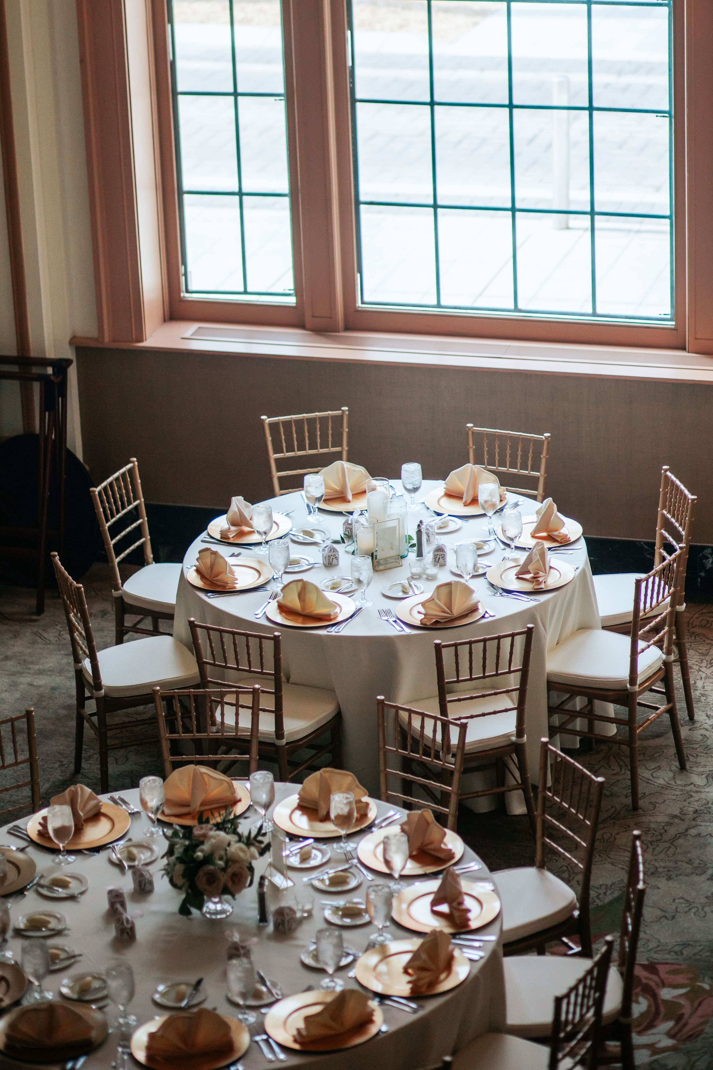 Round banquet tables set with white linens, elegant place settings and centerpieces in a bright windowed dining room.