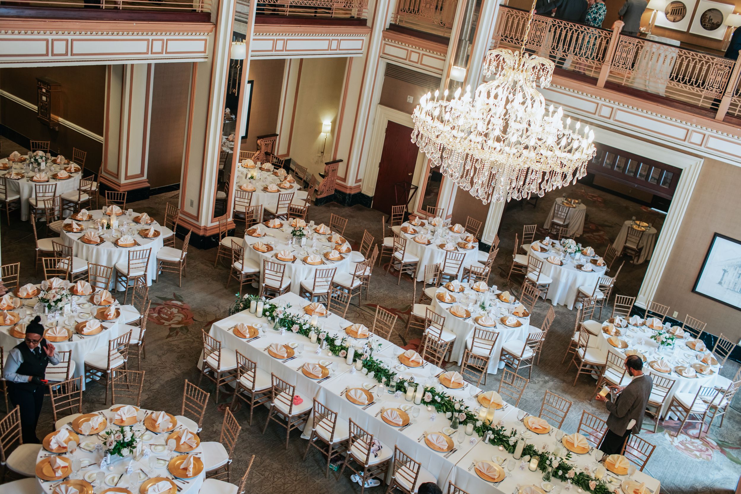 Elegant wedding reception ballroom with white tablecloths, crystal chandeliers and floral centerpieces arranged in rows.