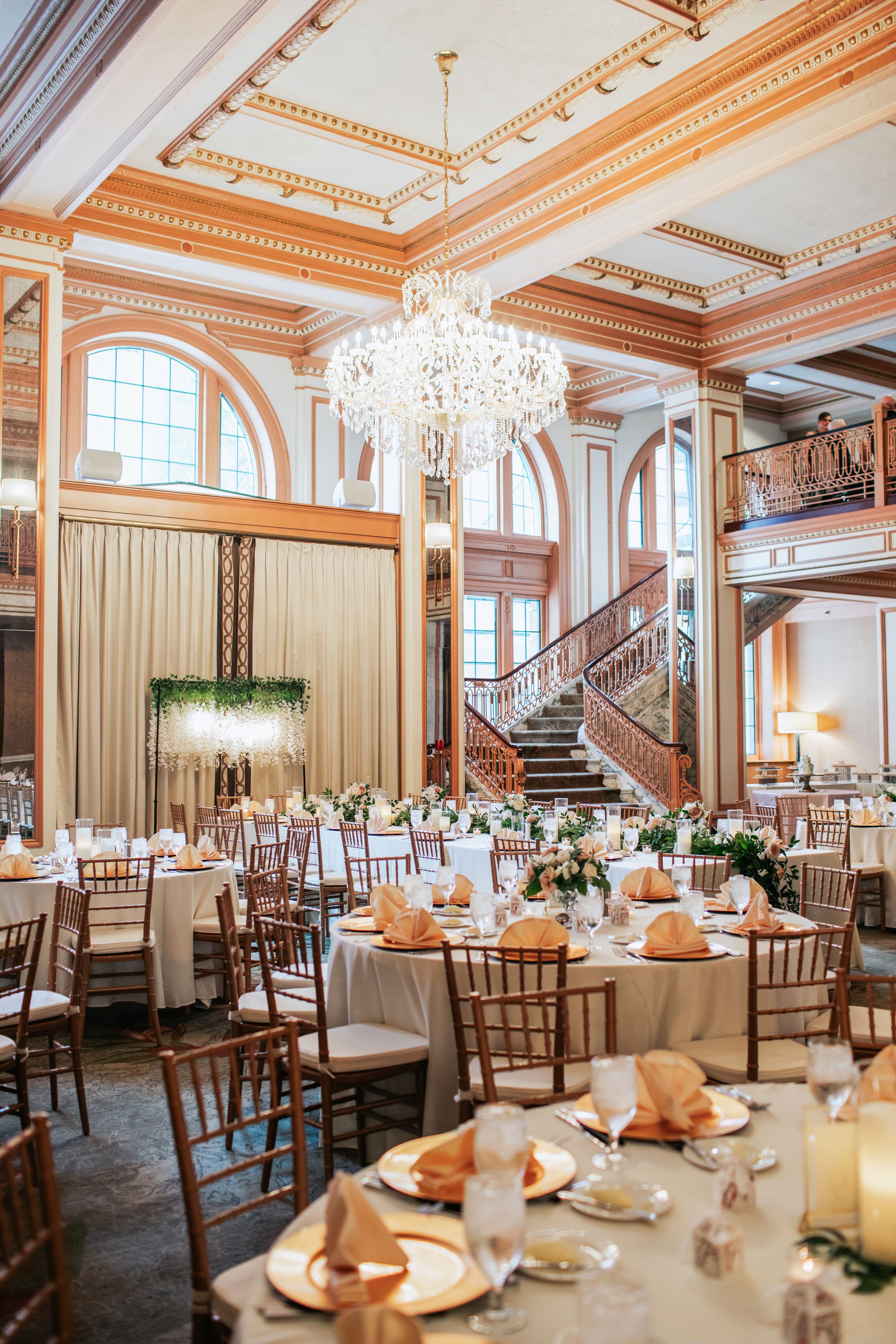 Elegant ballroom wedding reception setup with crystal chandelier, grand staircase, and peach-toned table settings.