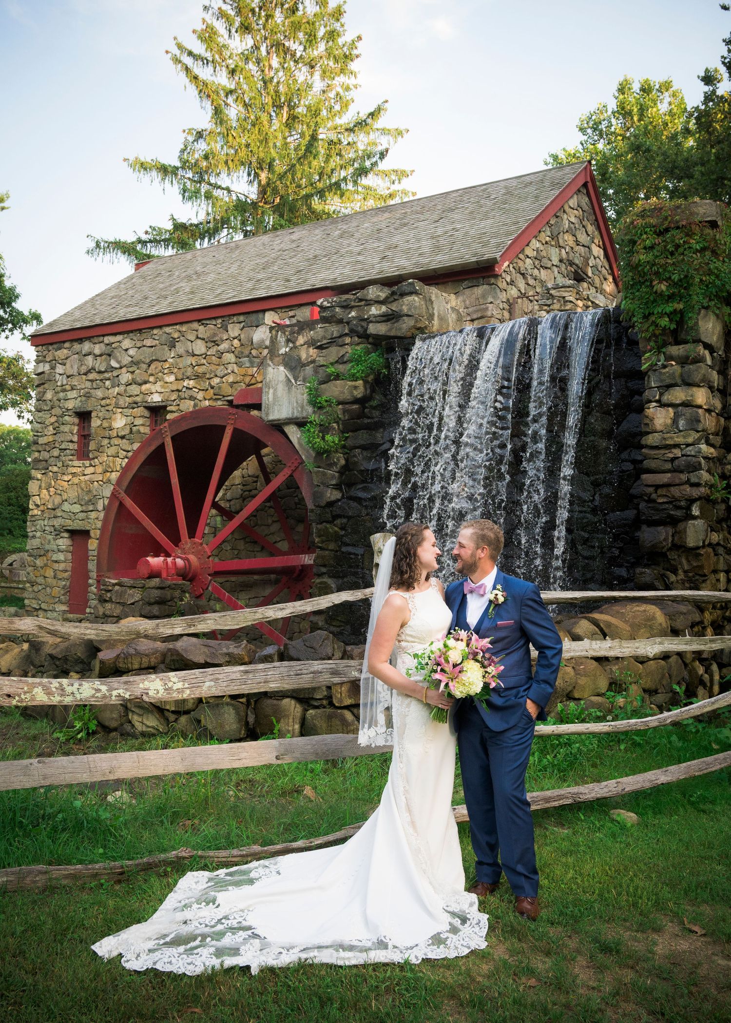 A historic stone mill with a red water wheel creates a romantic backdrop near a cascading waterfall.