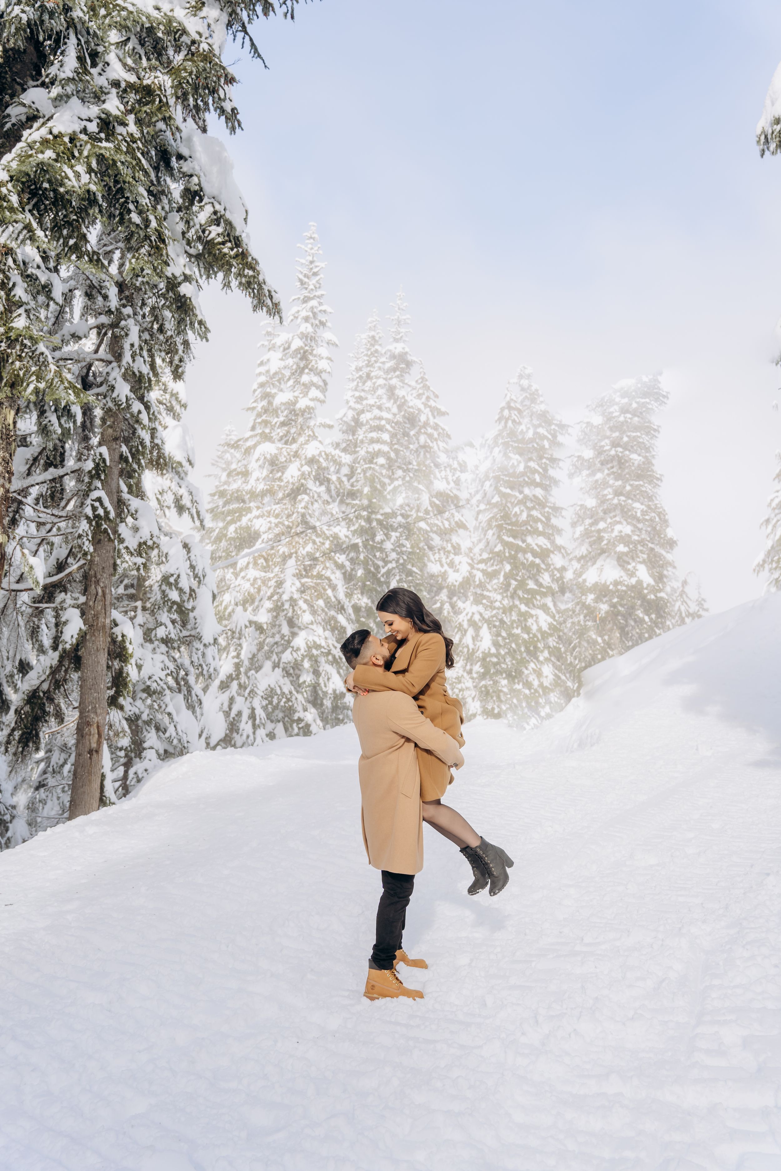 A couple embraces in a romantic moment while standing in deep snow with snow-covered evergreen trees in the background.