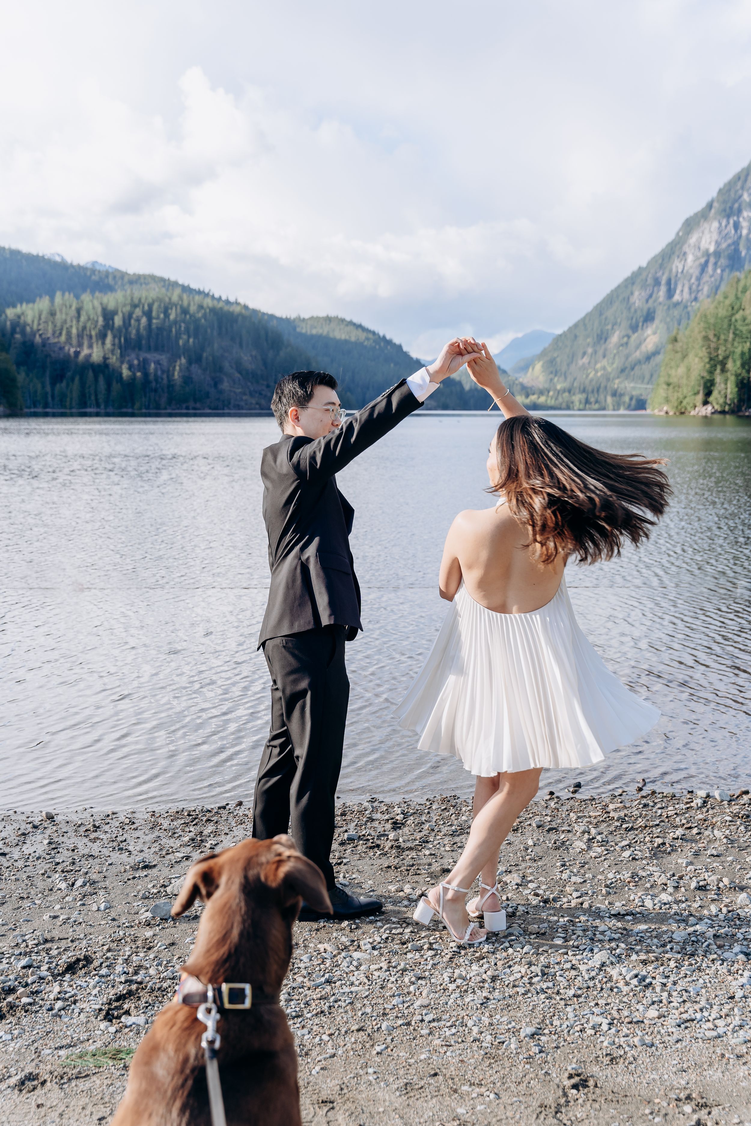A couple dances together on a rocky lakeshore with mountain views and their dog watching from the foreground.