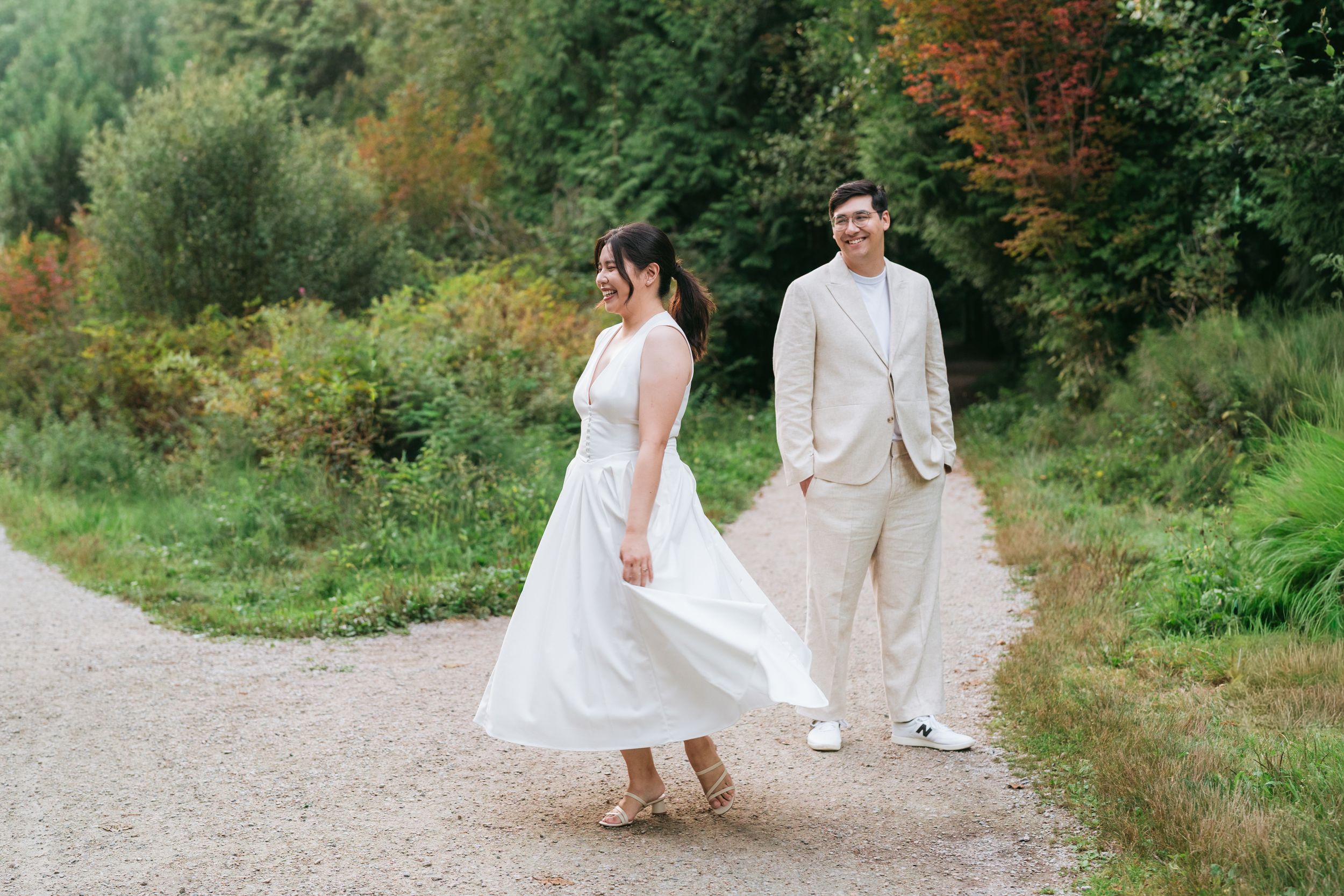 A couple in wedding attire walks along a scenic nature path surrounded by lush greenery and autumn foliage.