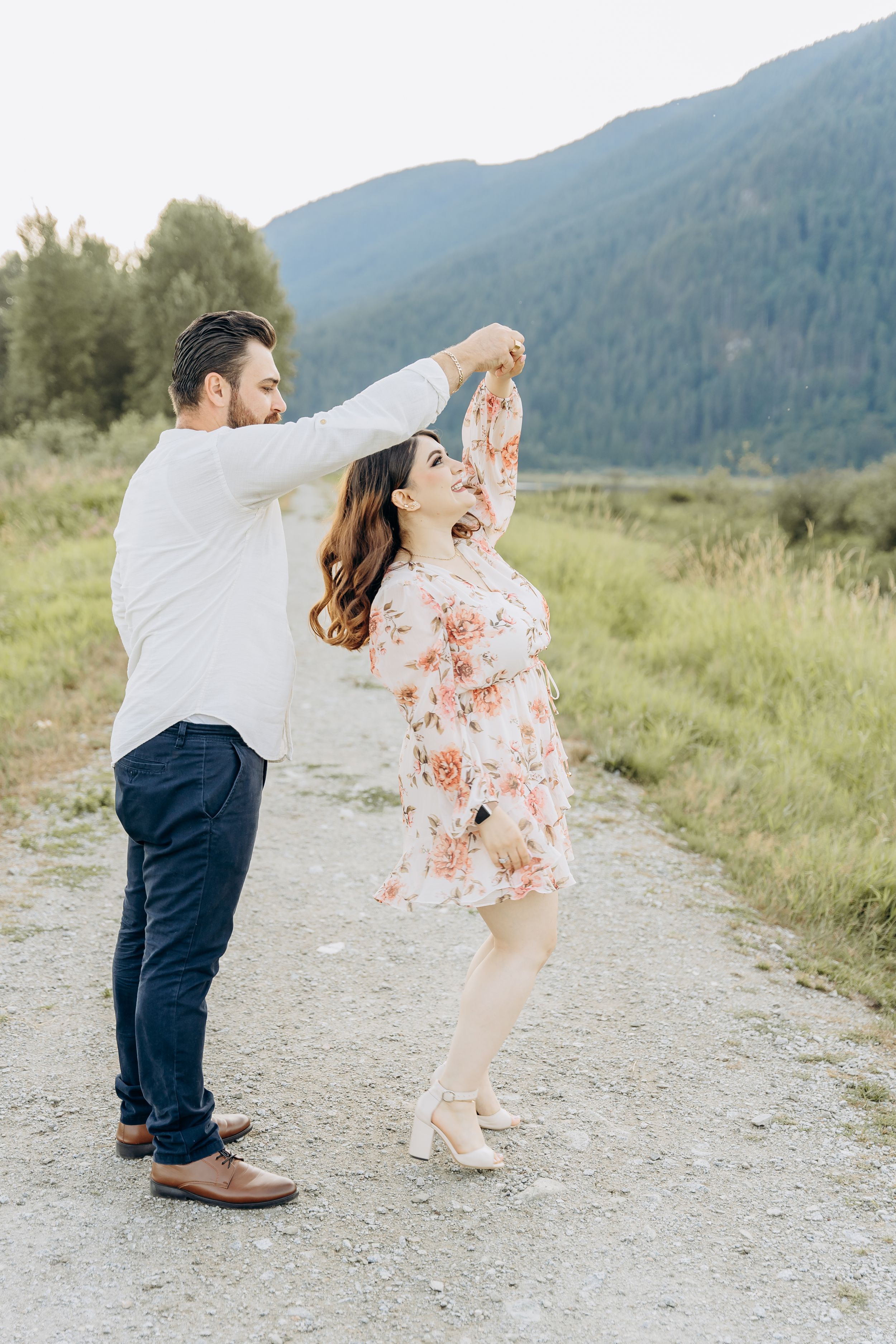 A couple dances playfully on a mountain trail surrounded by green meadows and hills.