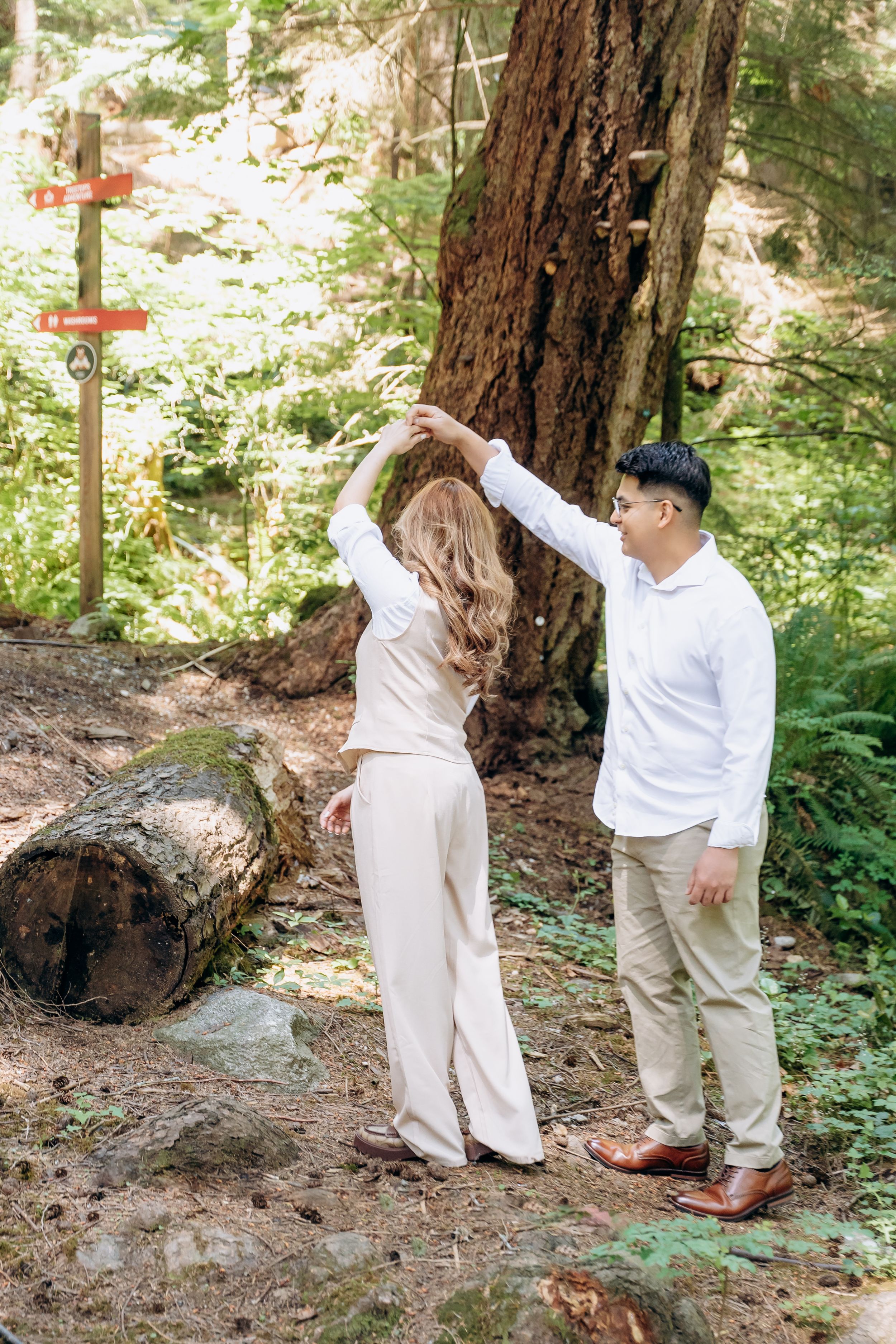 Two people share a tender moment among tall redwood trees in a sunlit forest setting.