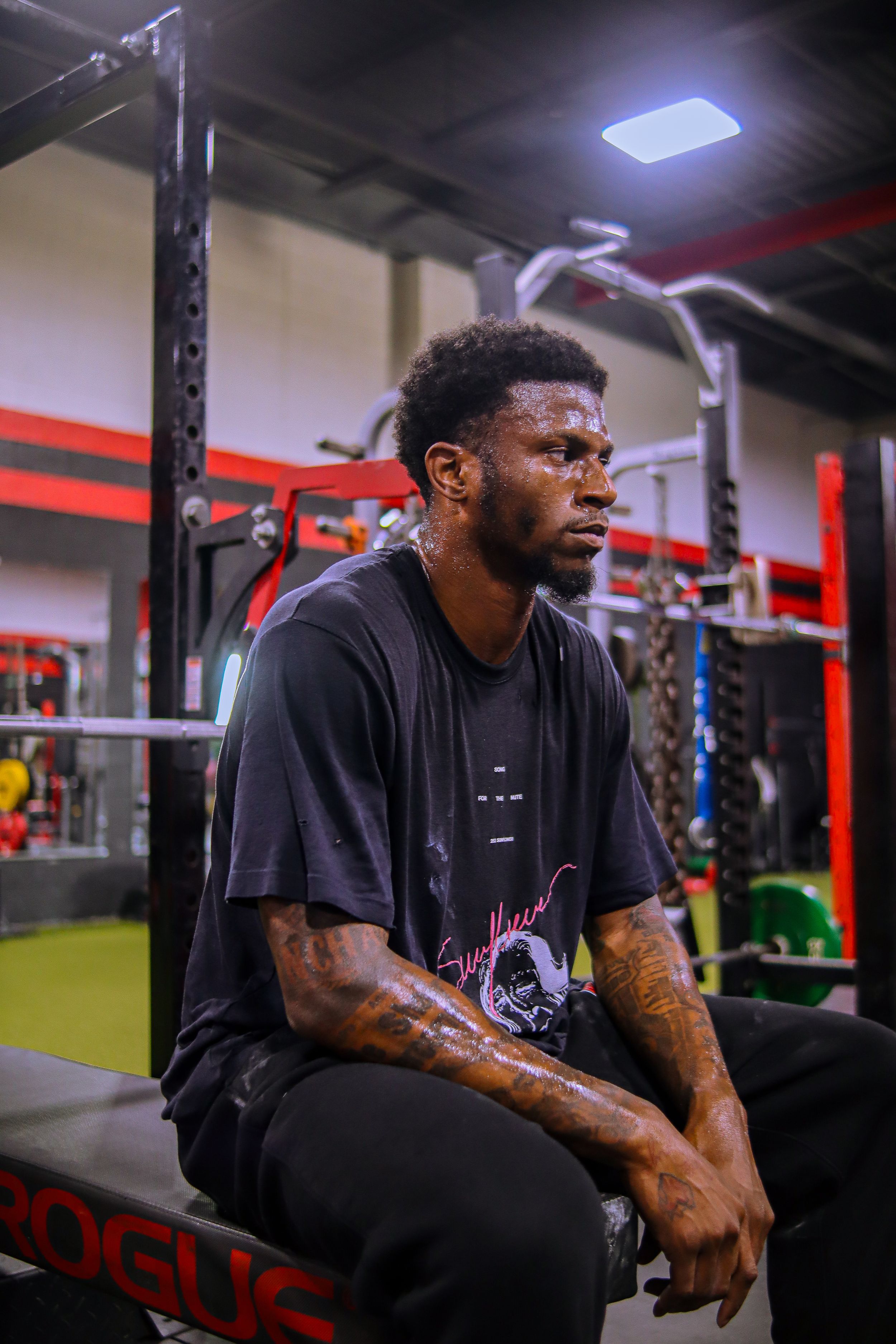 Tattooed athlete in black t-shirt resting on Rogue bench in red and black gym after workout.