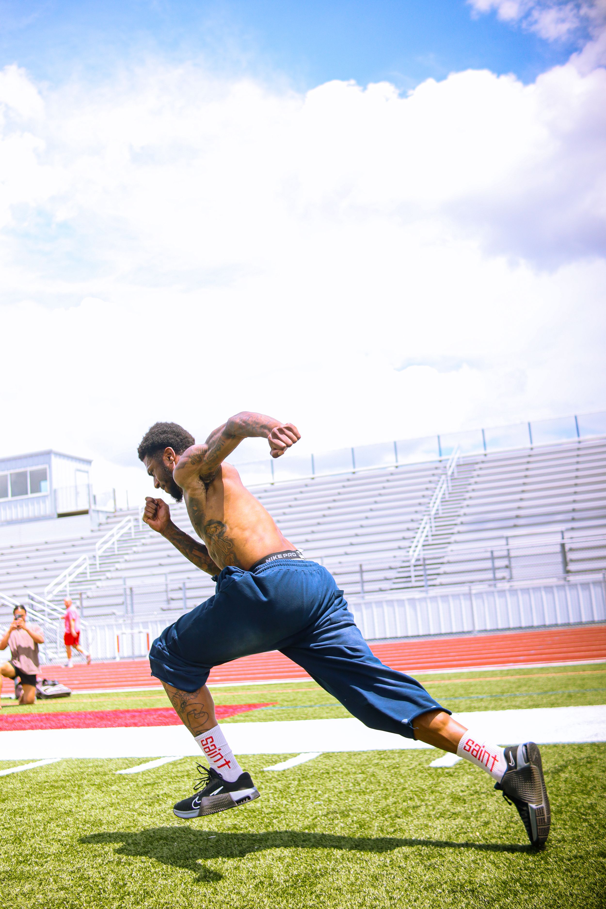 Shirtless athlete sprinting on green turf field at stadium, wearing blue shorts and white socks, training outdoors.