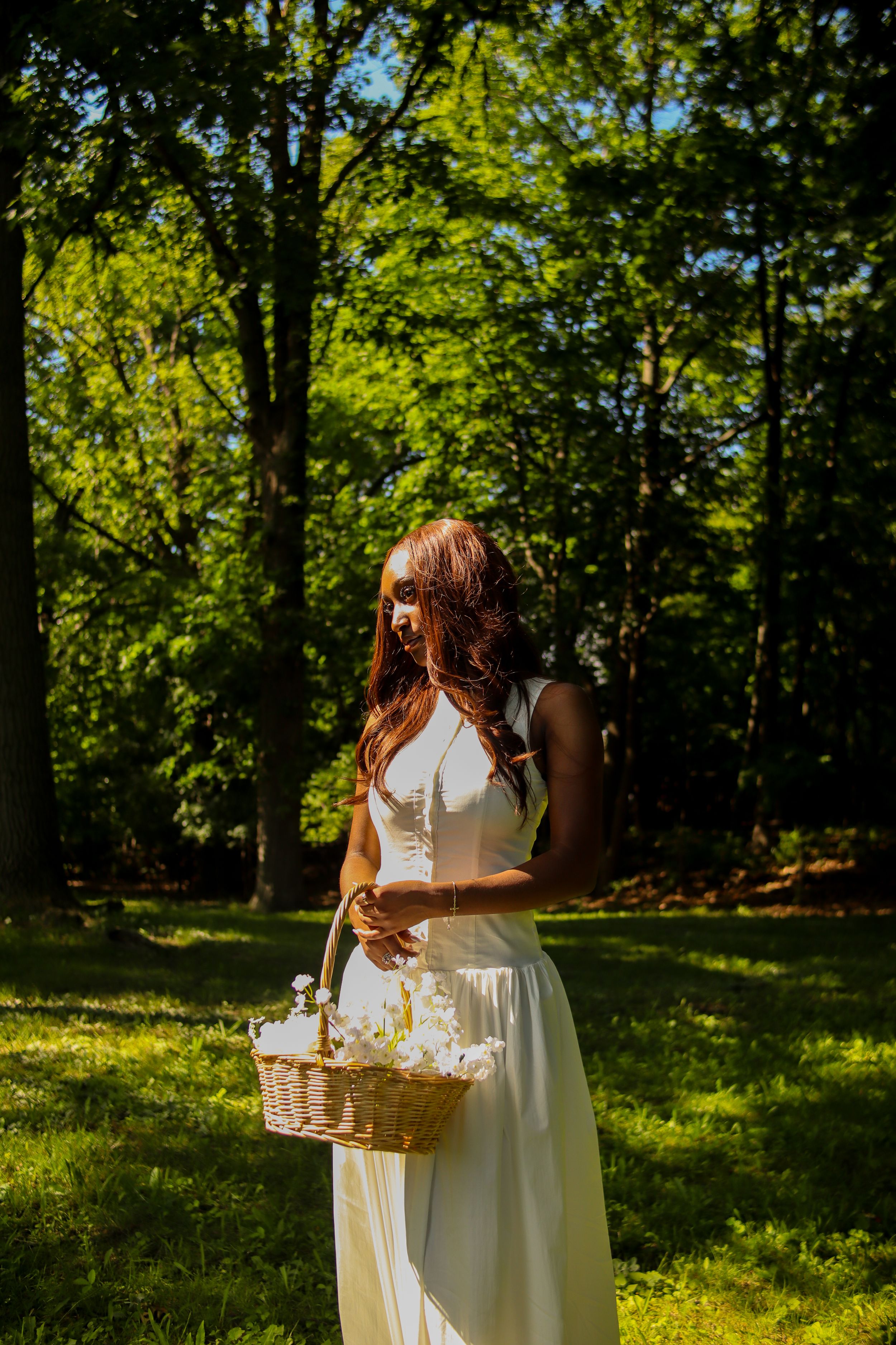Woman in white dress holding a wicker basket with flowers, standing in a sunlit green park surrounded by trees.