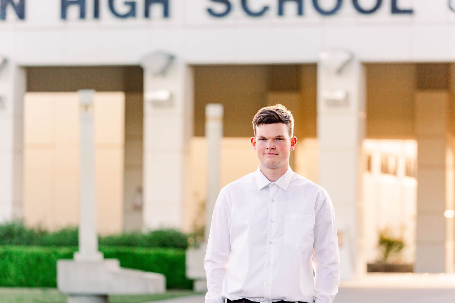 A student in a white button-down shirt stands in front of a high school building during golden hour.
