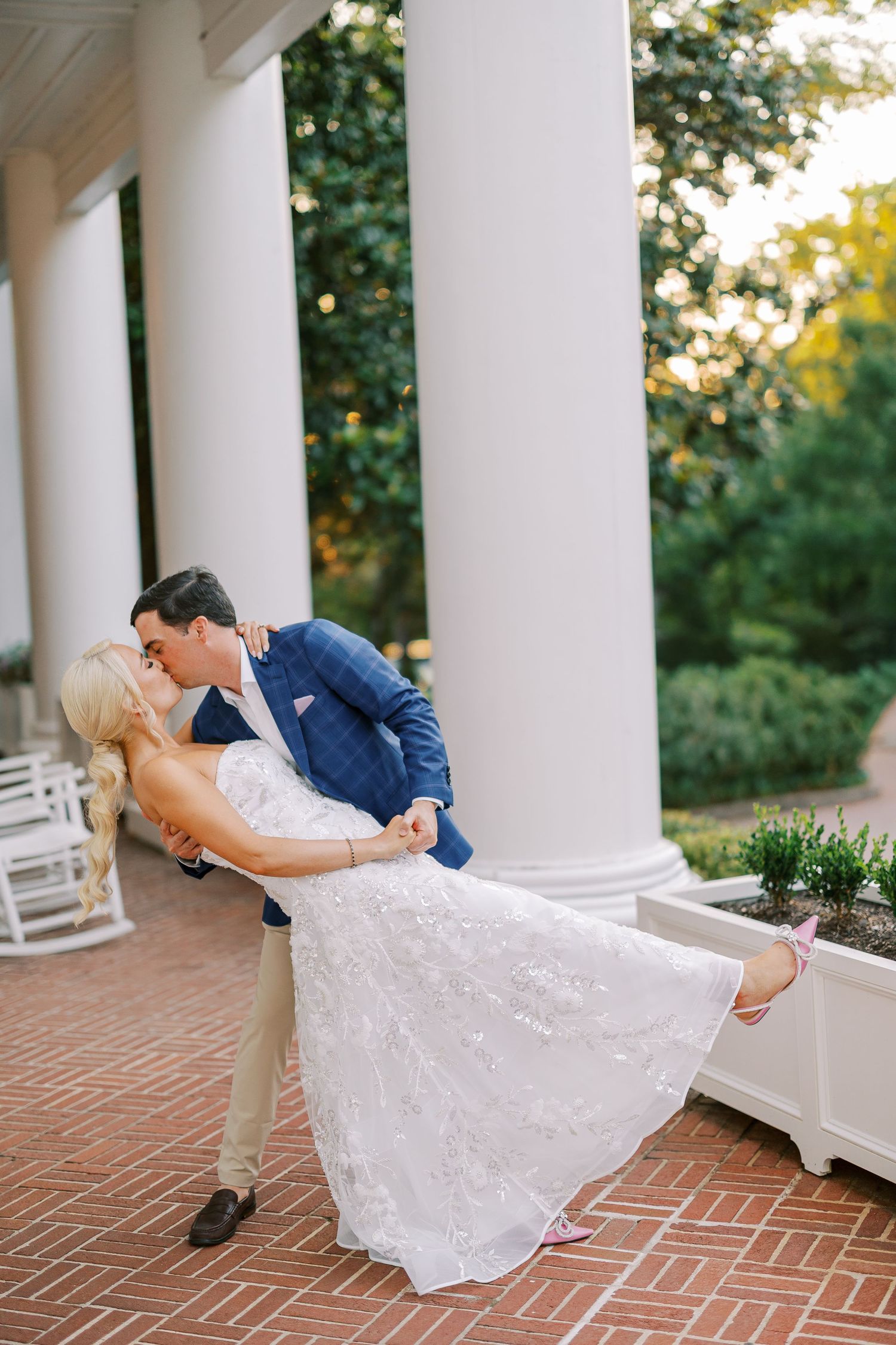 A romantic couple in wedding attire share a dip kiss on an elegant white-columned porch with brick flooring.