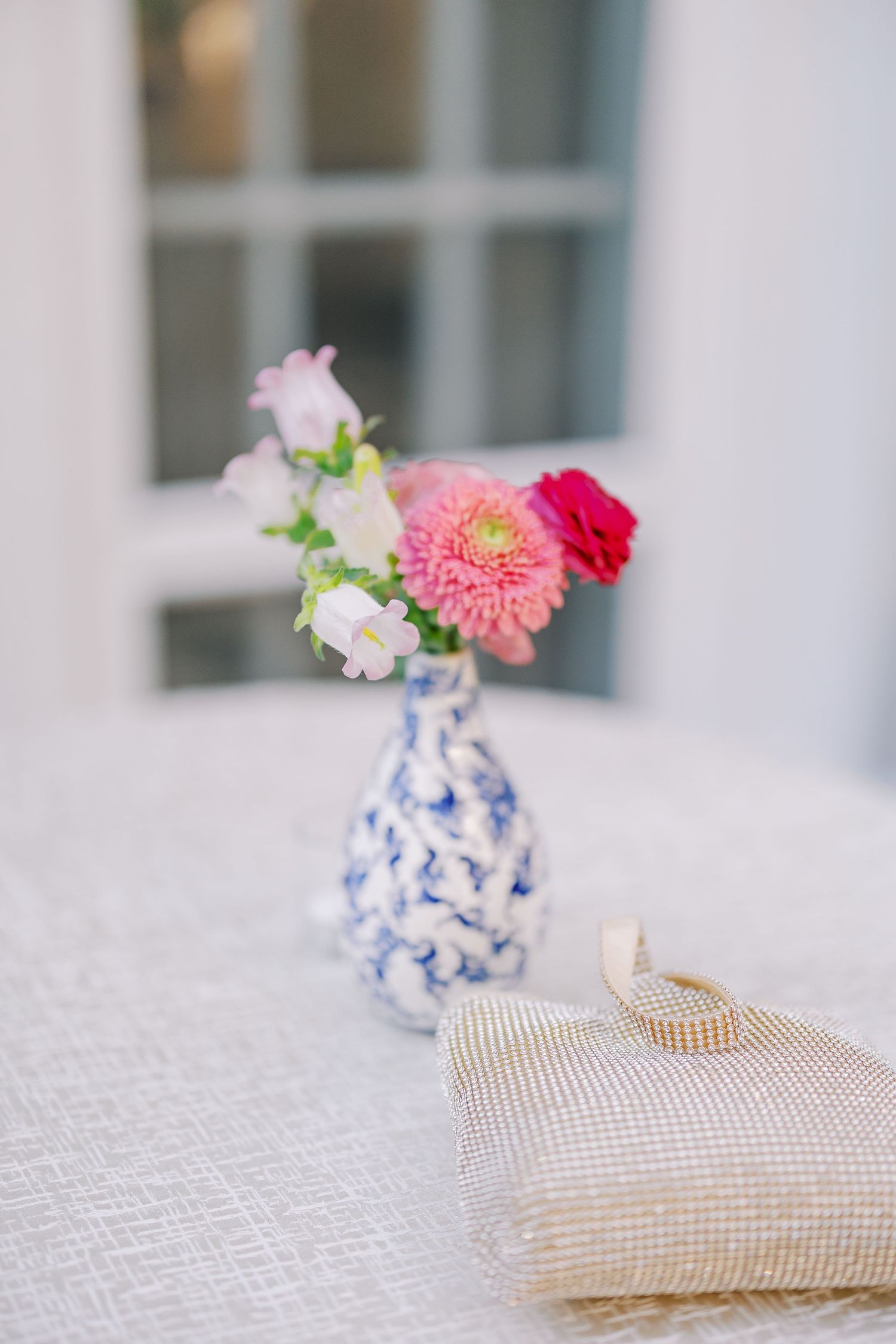 Wedding rings rest on a textured pillow beside a blue and white vase with pink and red flowers.