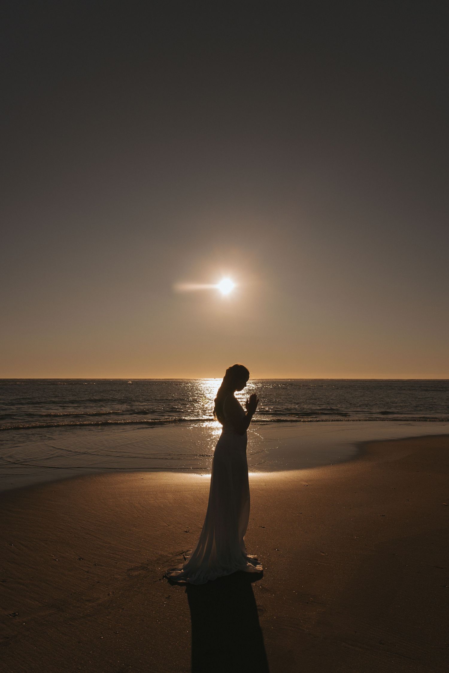 Woman in white dress silhouetted against golden sunset on sandy beach near ocean waves in New Zealand.