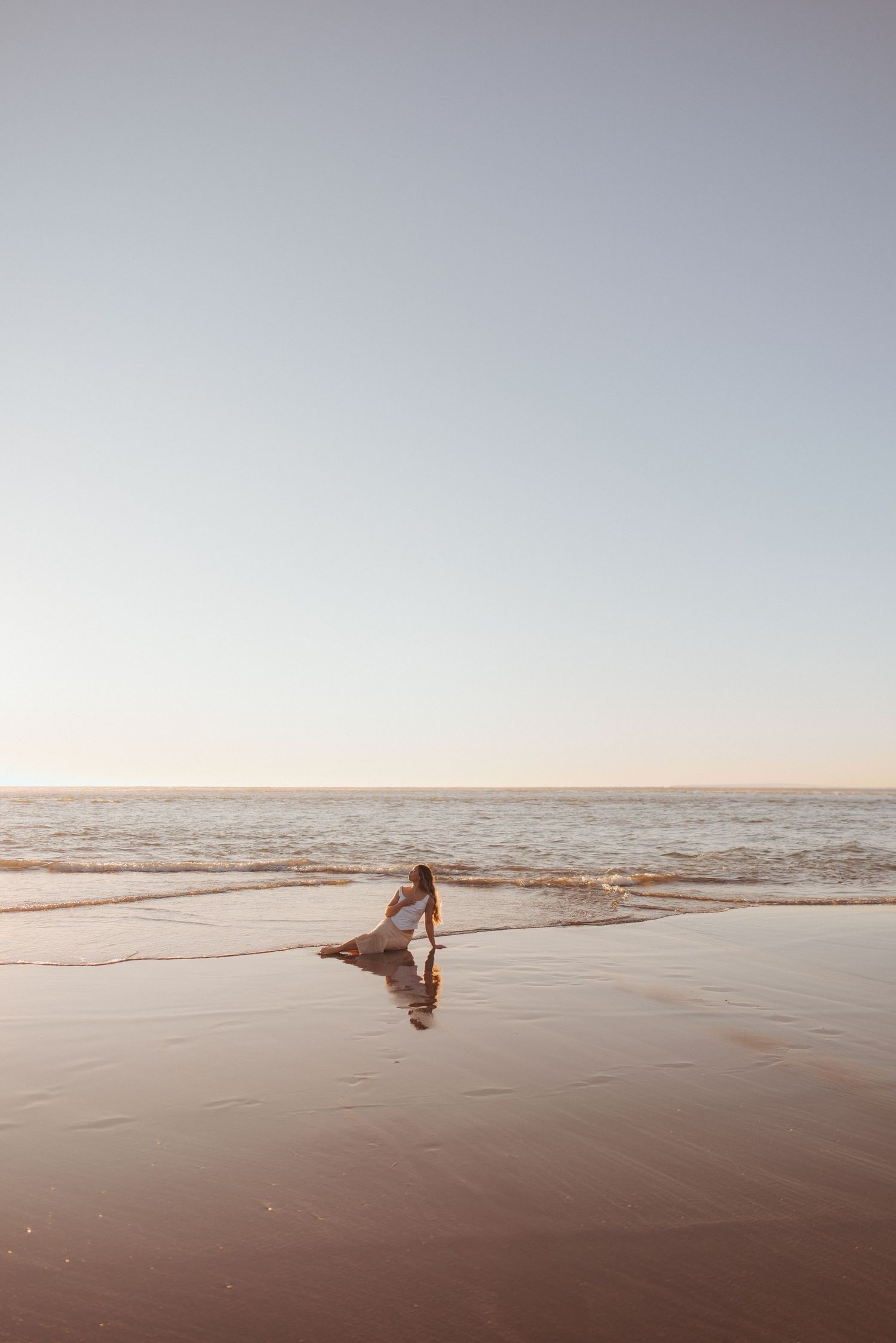 Woman in white dress walking along wet sandy beach shore at golden hour sunset in New Zealand.
