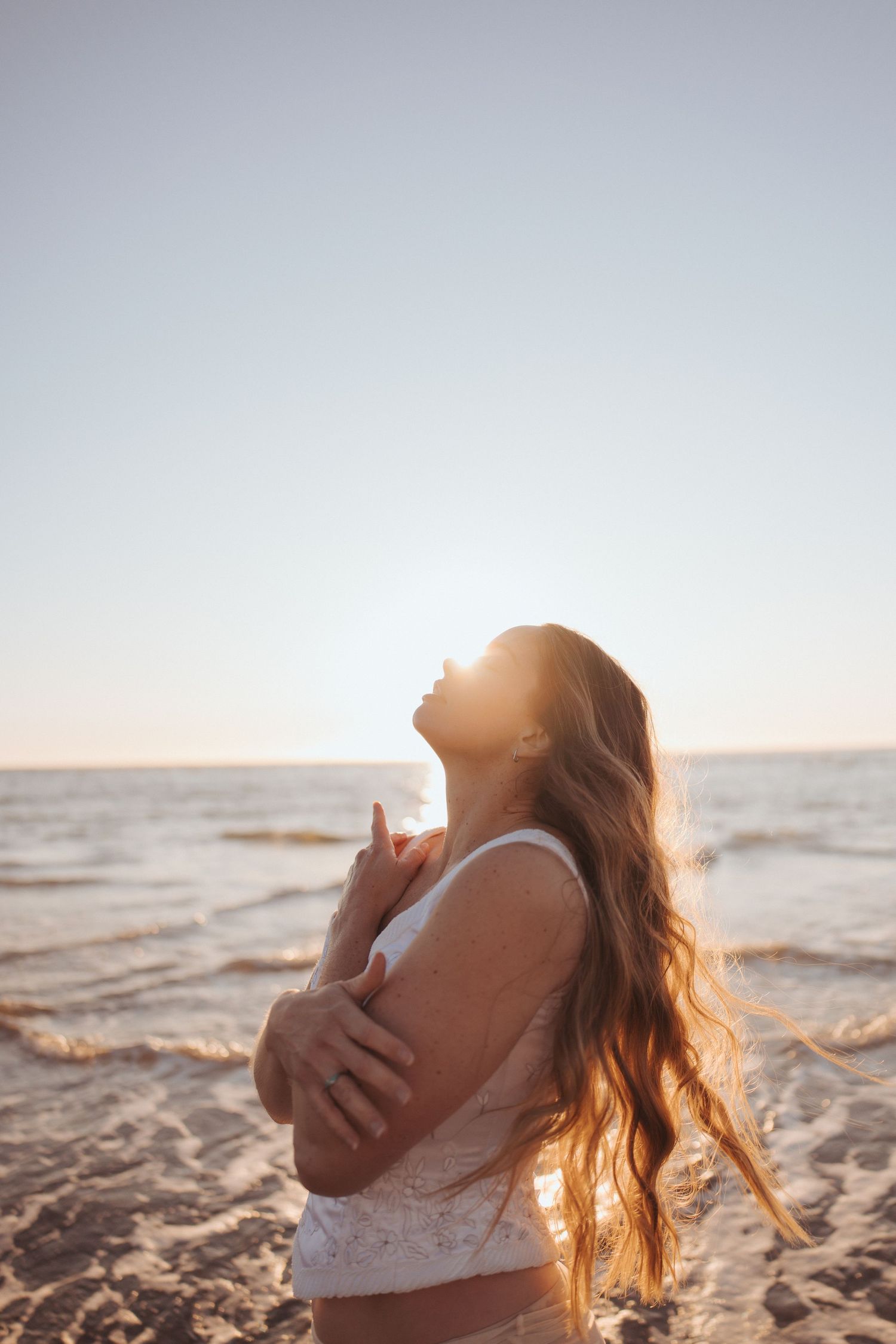 Woman with long hair embracing herself on sunny beach with ocean waves in background in New Zealand.