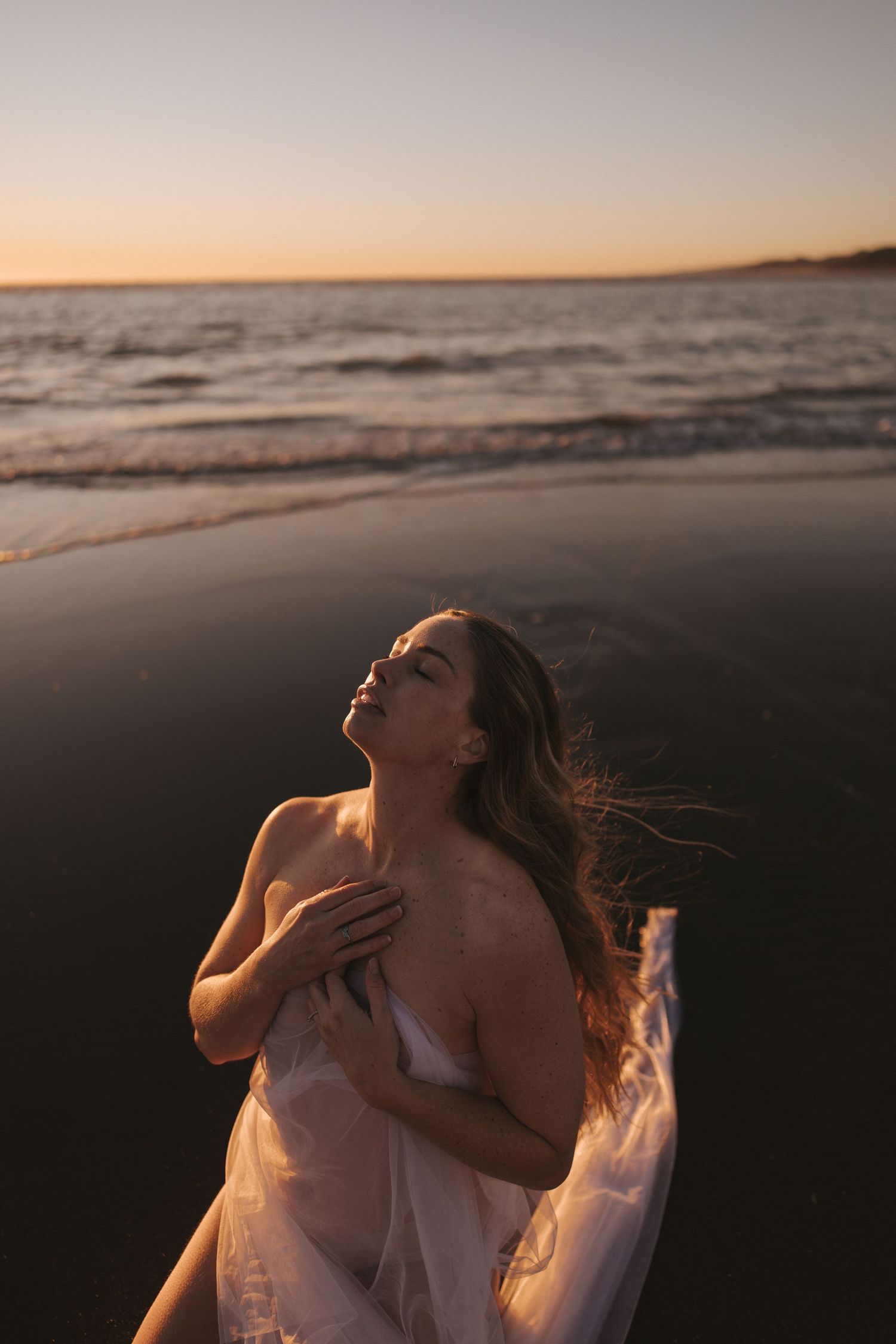 Woman in flowing white dress holding fabric on dark beach at dramatic sunset in New Zealand.