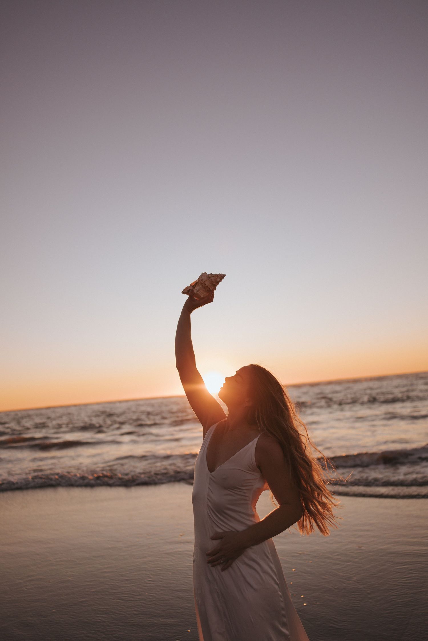 Woman in white dress raising arm toward sunset sky on beach with ocean behind her in New Zealand.