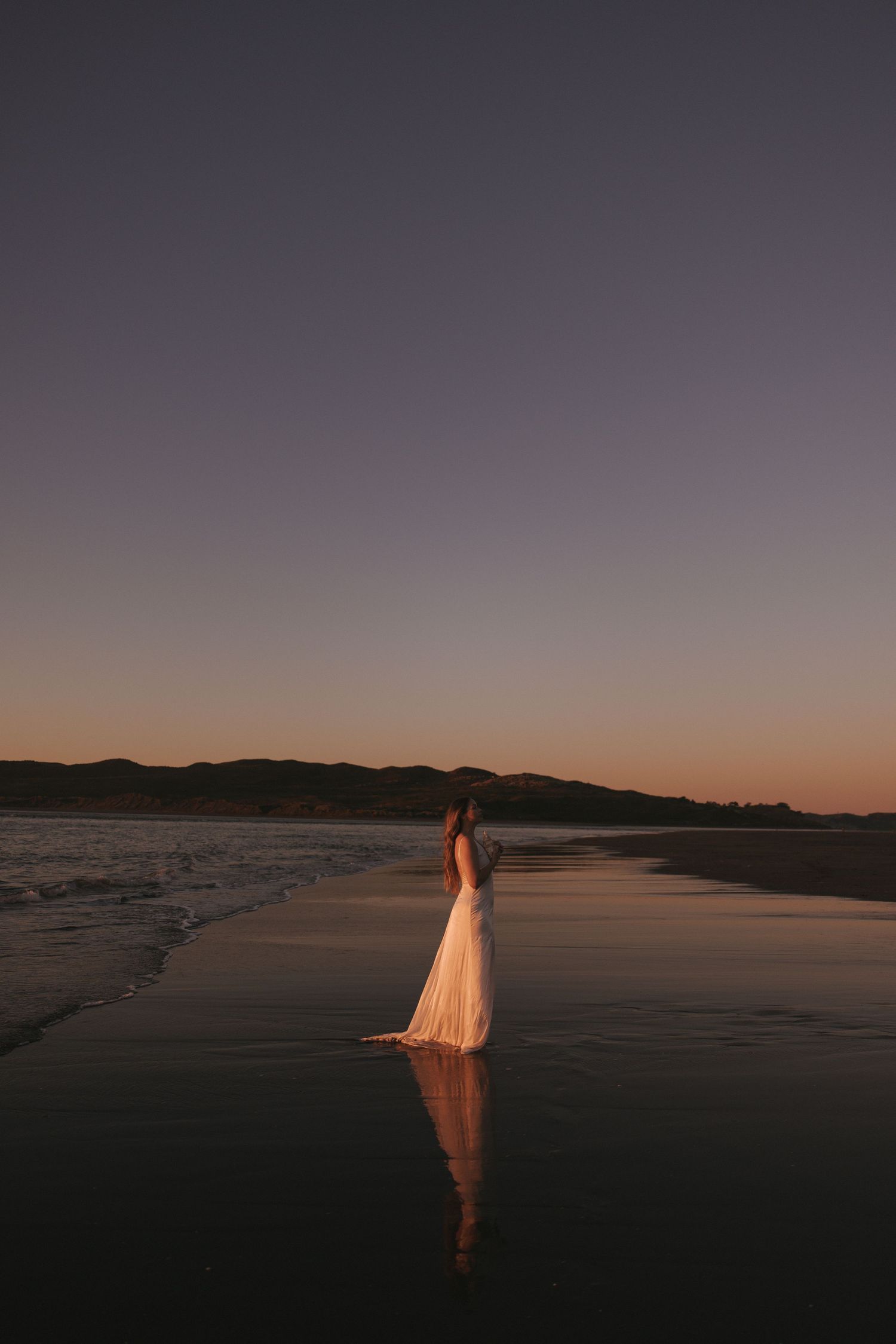Woman in elegant white flowing dress standing on reflective beach at twilight sunset in New Zealand.
