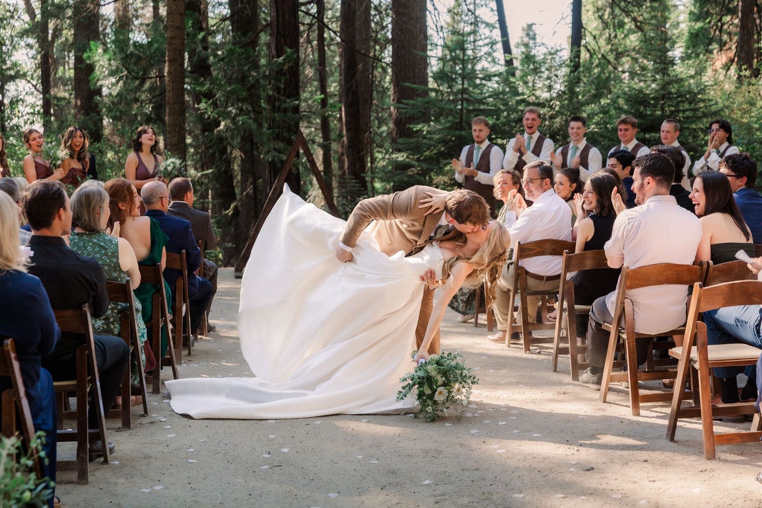 A couple shares their first dance during an outdoor forest wedding ceremony surrounded by wooden chairs and guests.