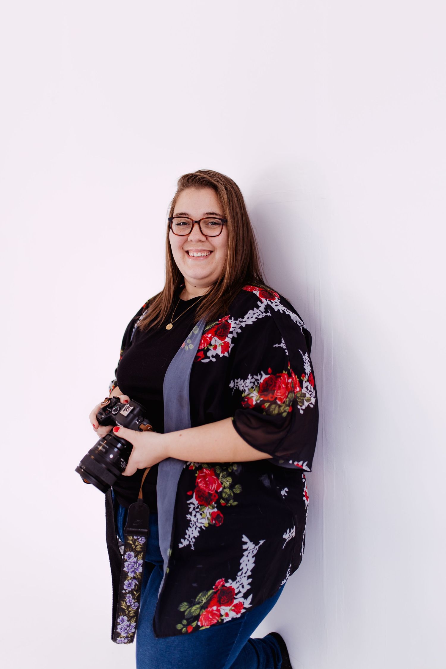 Smiling female photographer holding a DSLR camera, wearing floral kimono and glasses against white wall.