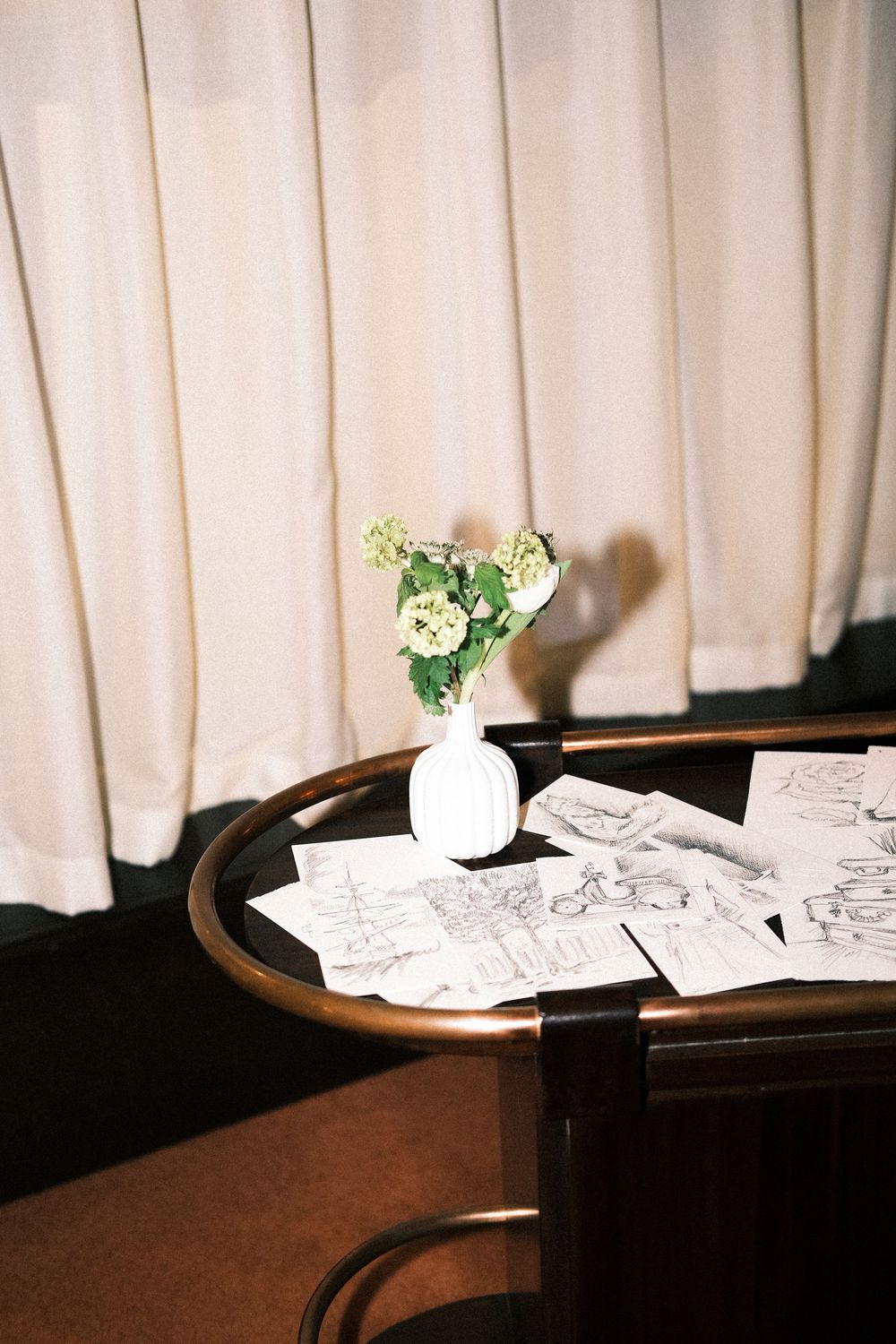 White flowers in a small vase sit atop a dark wooden table with papers scattered around it.
