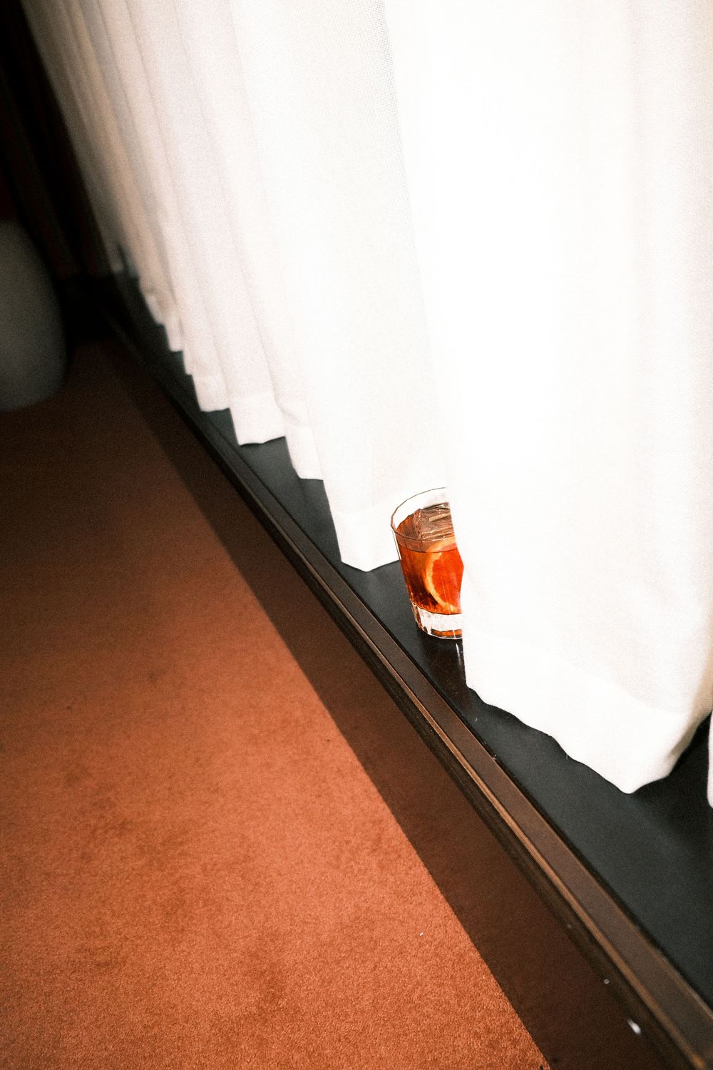 A glass of orange liquid sits on a windowsill between white curtains and red carpet.