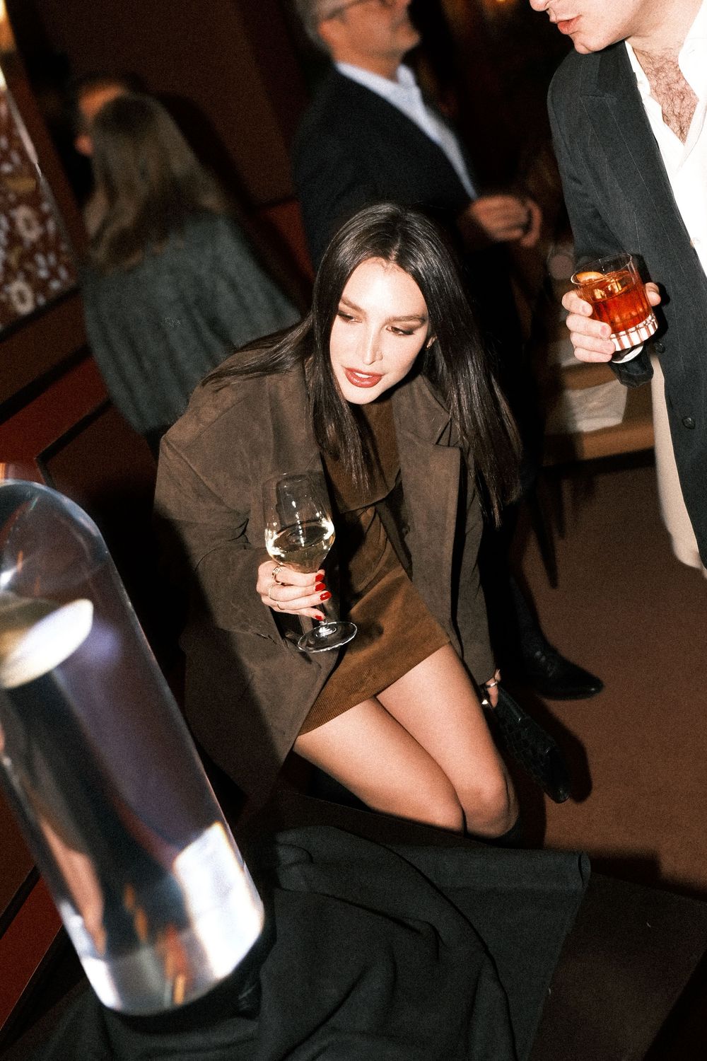 A person in a black outfit holds a glass at an evening social event.