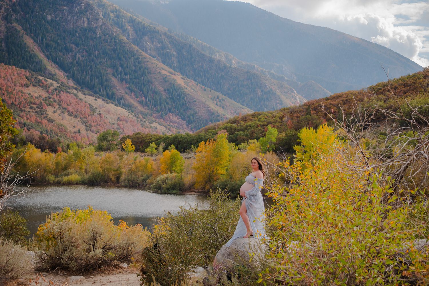 Woman in white dress standing near lake surrounded by colorful autumn foliage and mountains.