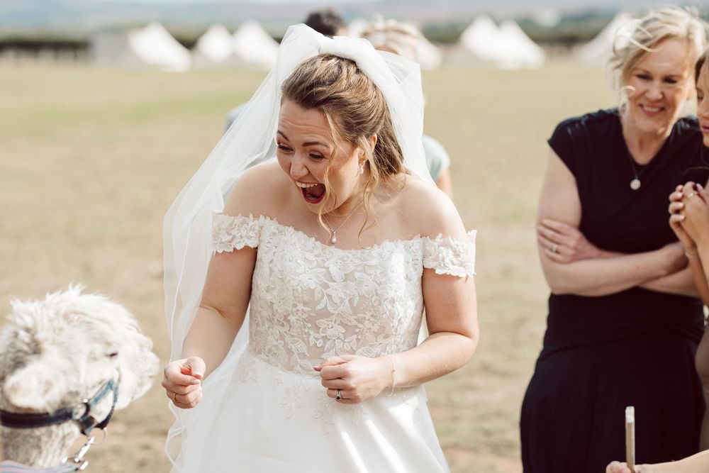 A bride in an off-shoulder lace wedding dress laughs while standing outdoors on a sunny day.