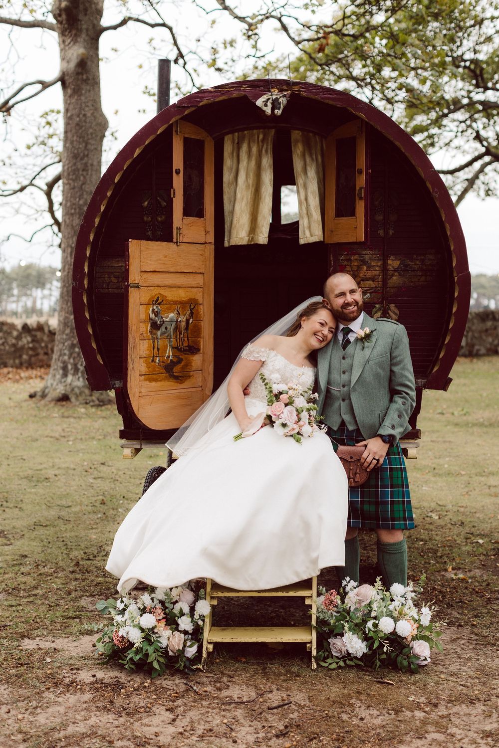 A romantic wedding couple poses in front of rustic wooden barrel-shaped glamping pods decorated with floral arrangements.
