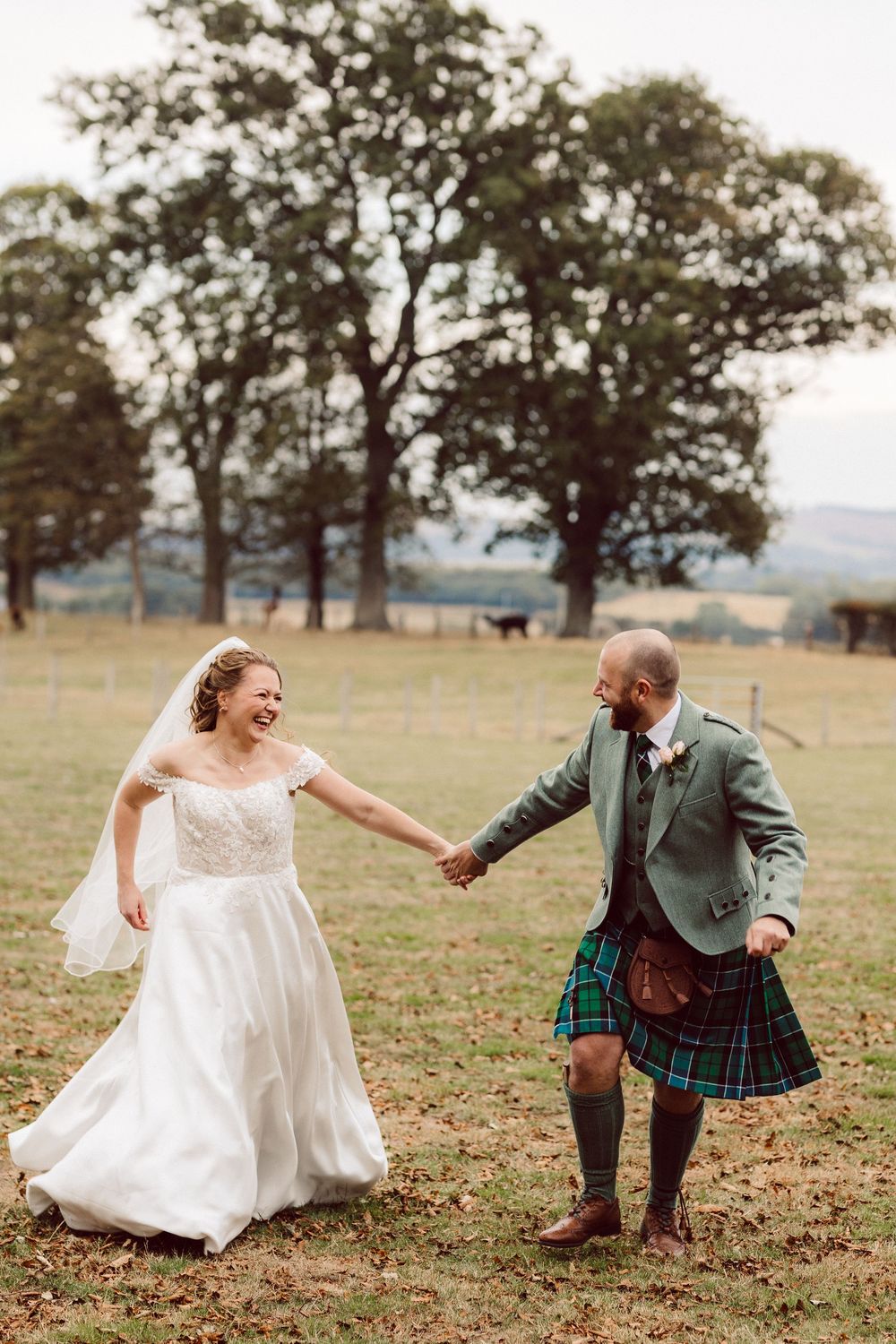 A couple in wedding attire playfully runs and dances together in a grassy field with trees in the background.