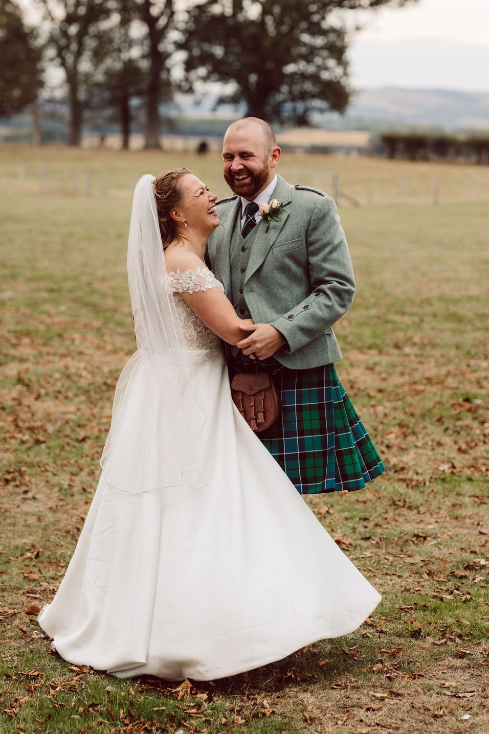 A couple in wedding attire dances outdoors, with the groom wearing a traditional green jacket and tartan kilt.