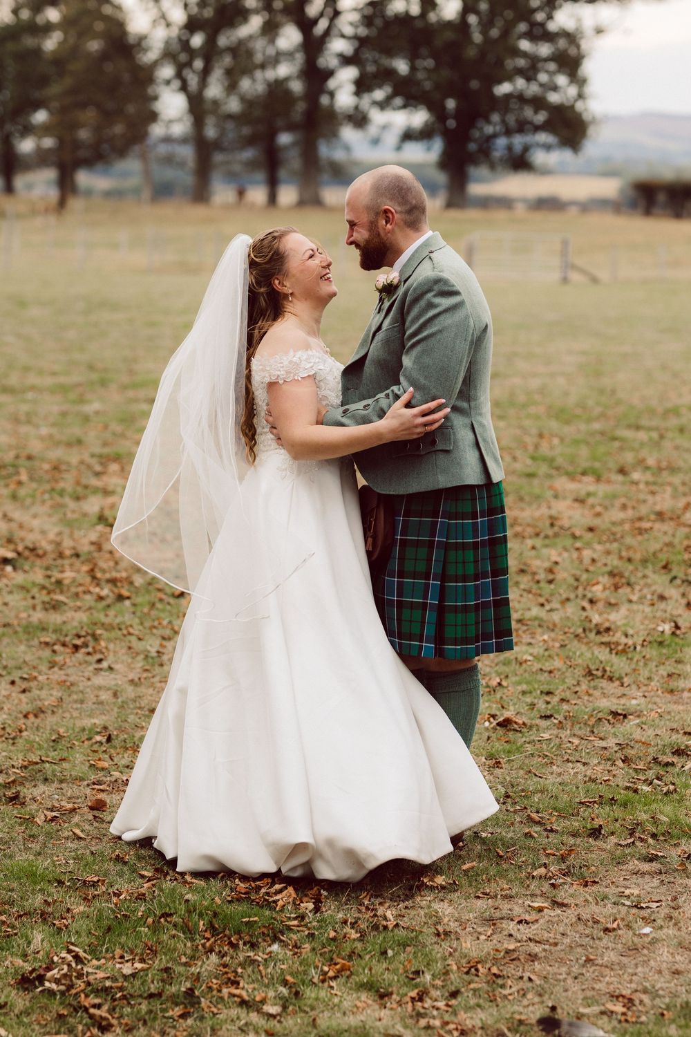 A couple in wedding attire shares a joyful moment outdoors, with the groom wearing a traditional Scottish kilt.