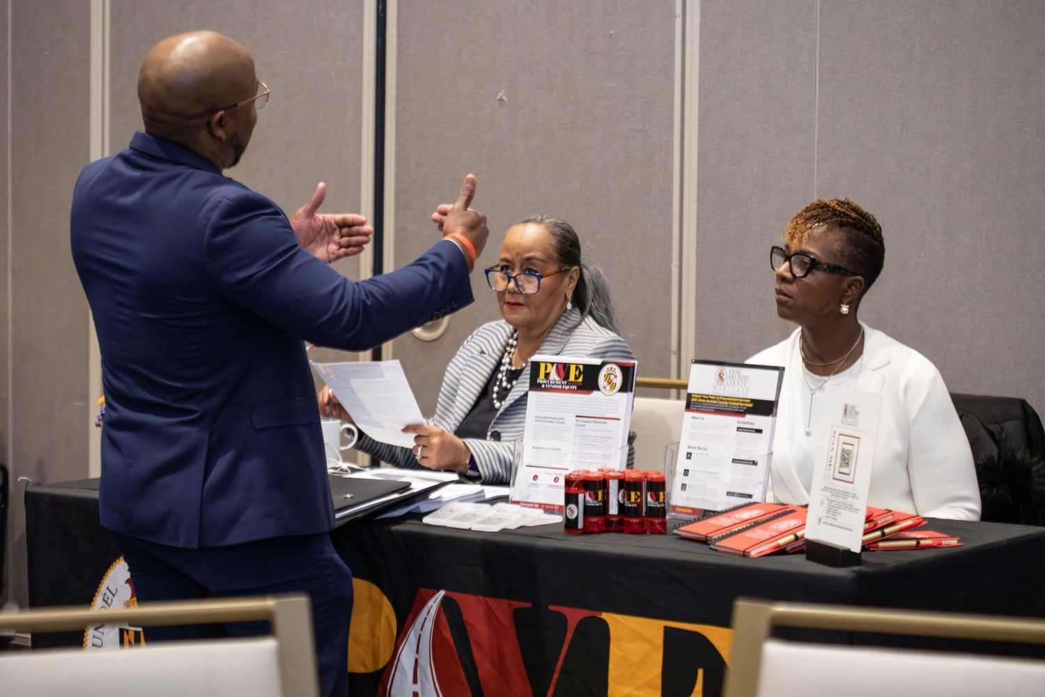 People engage in animated discussion at a business meeting table with Maryland flag decor.
