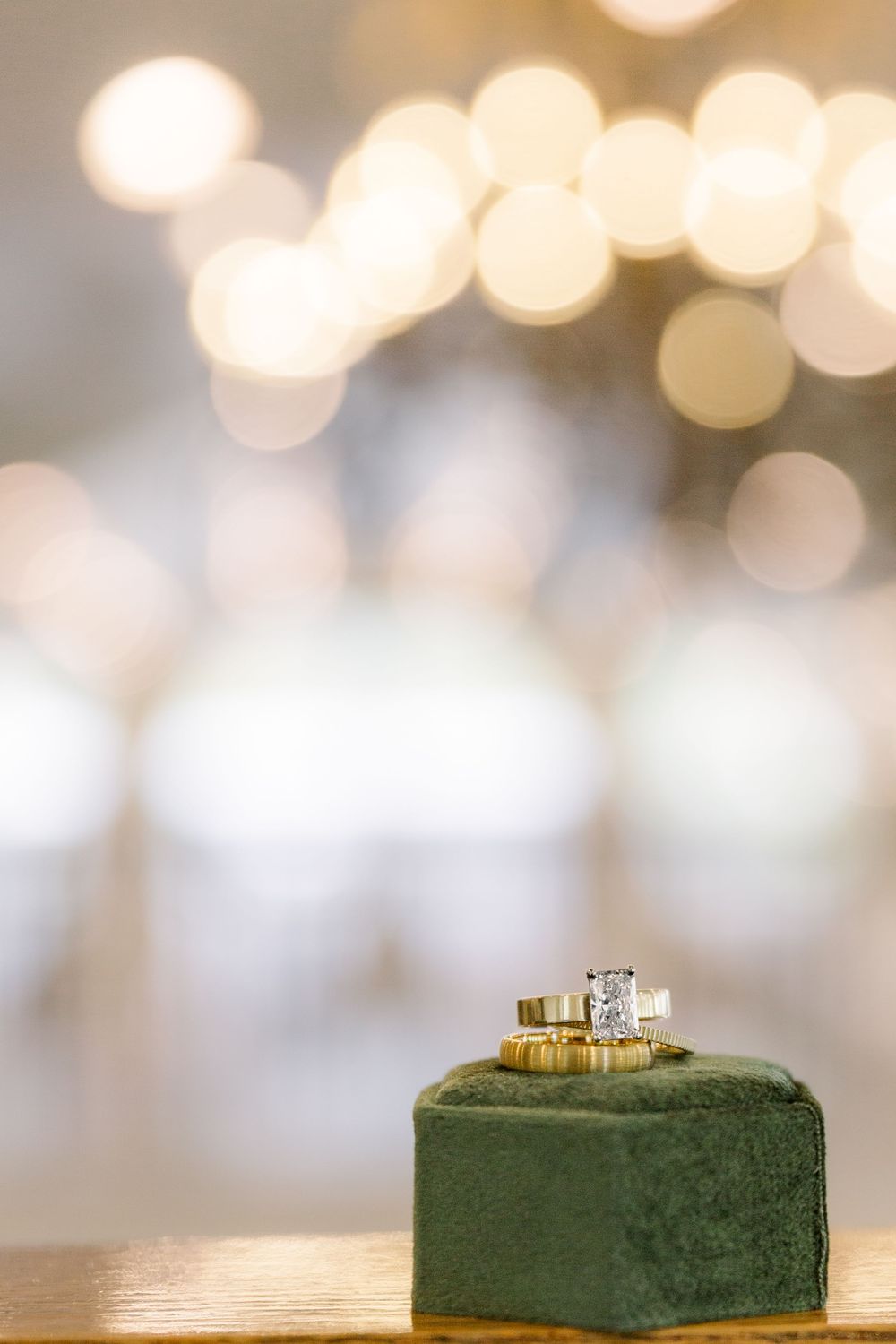 Two diamond engagement rings sit on a green velvet cushion with soft bokeh lights in the background.
