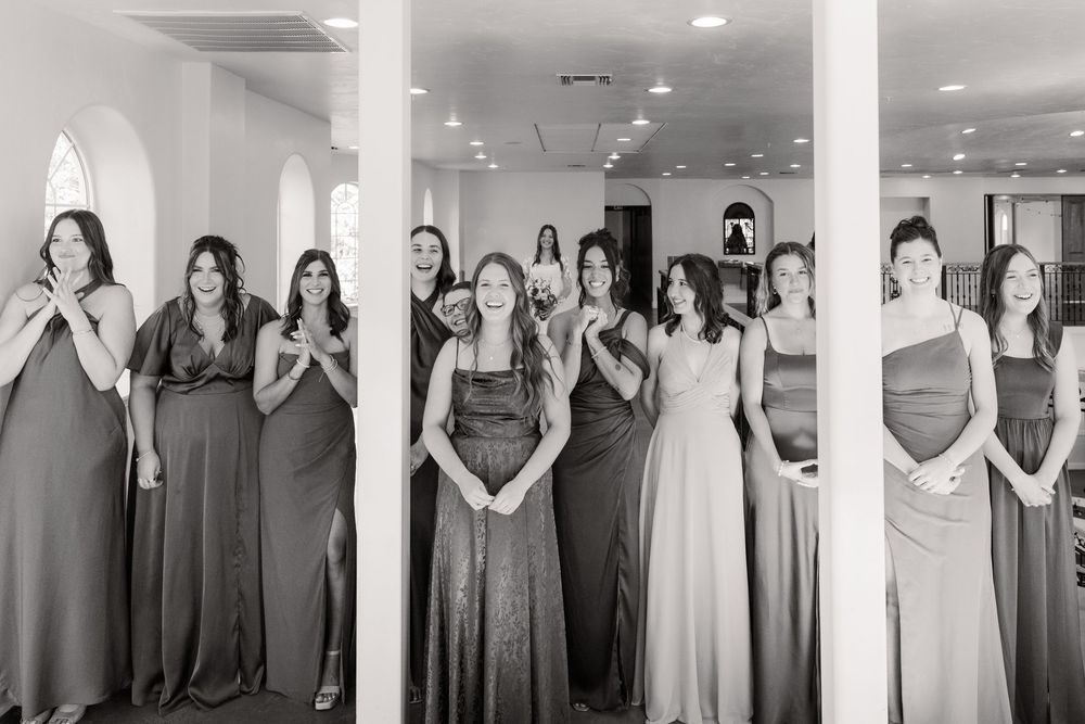 A group of bridesmaids in various shades of dresses stand together in an indoor venue during a wedding celebration.
