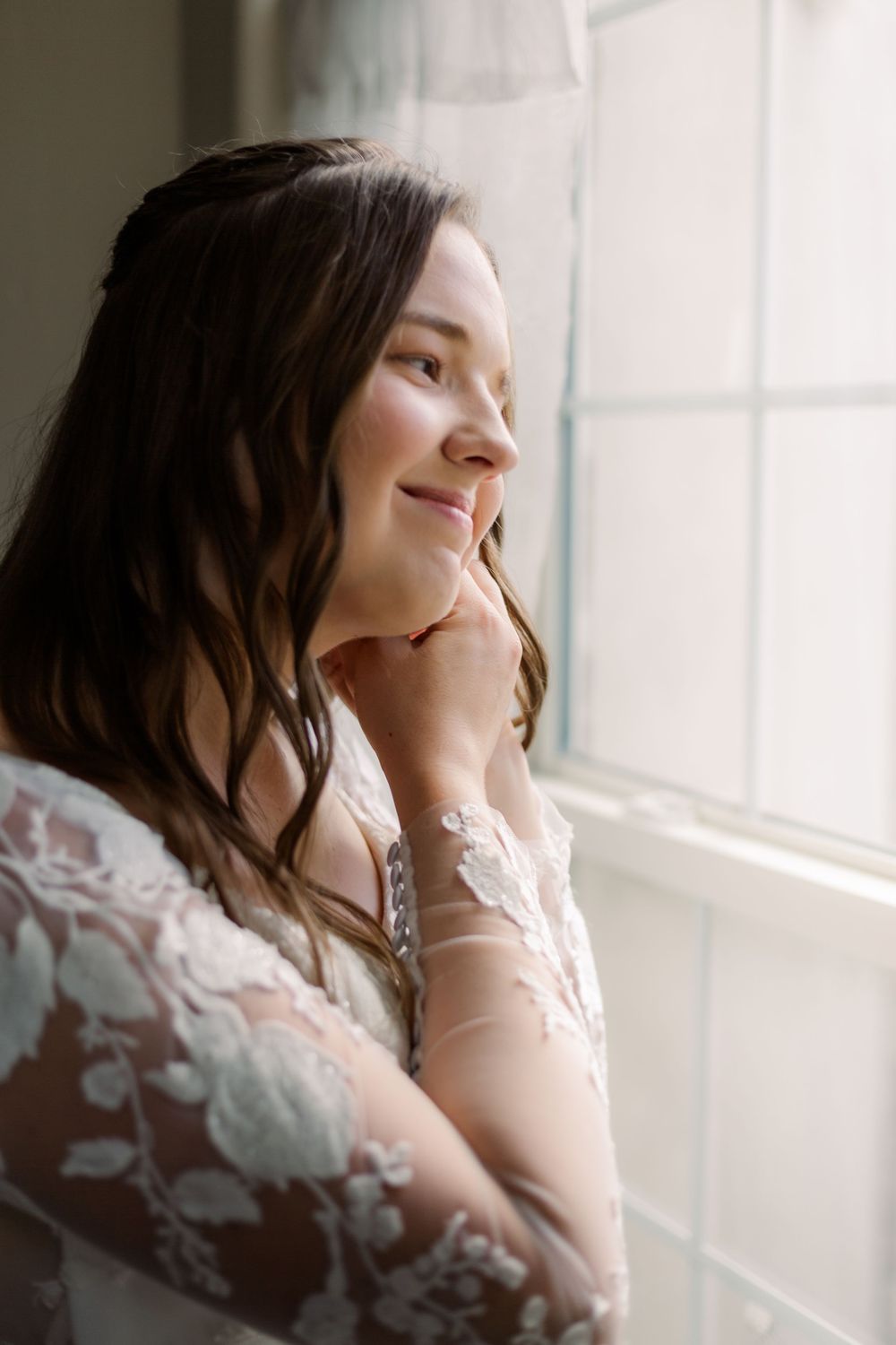 A dreamy portrait of someone in a delicate lace wedding dress gazing thoughtfully through a window in soft natural light.