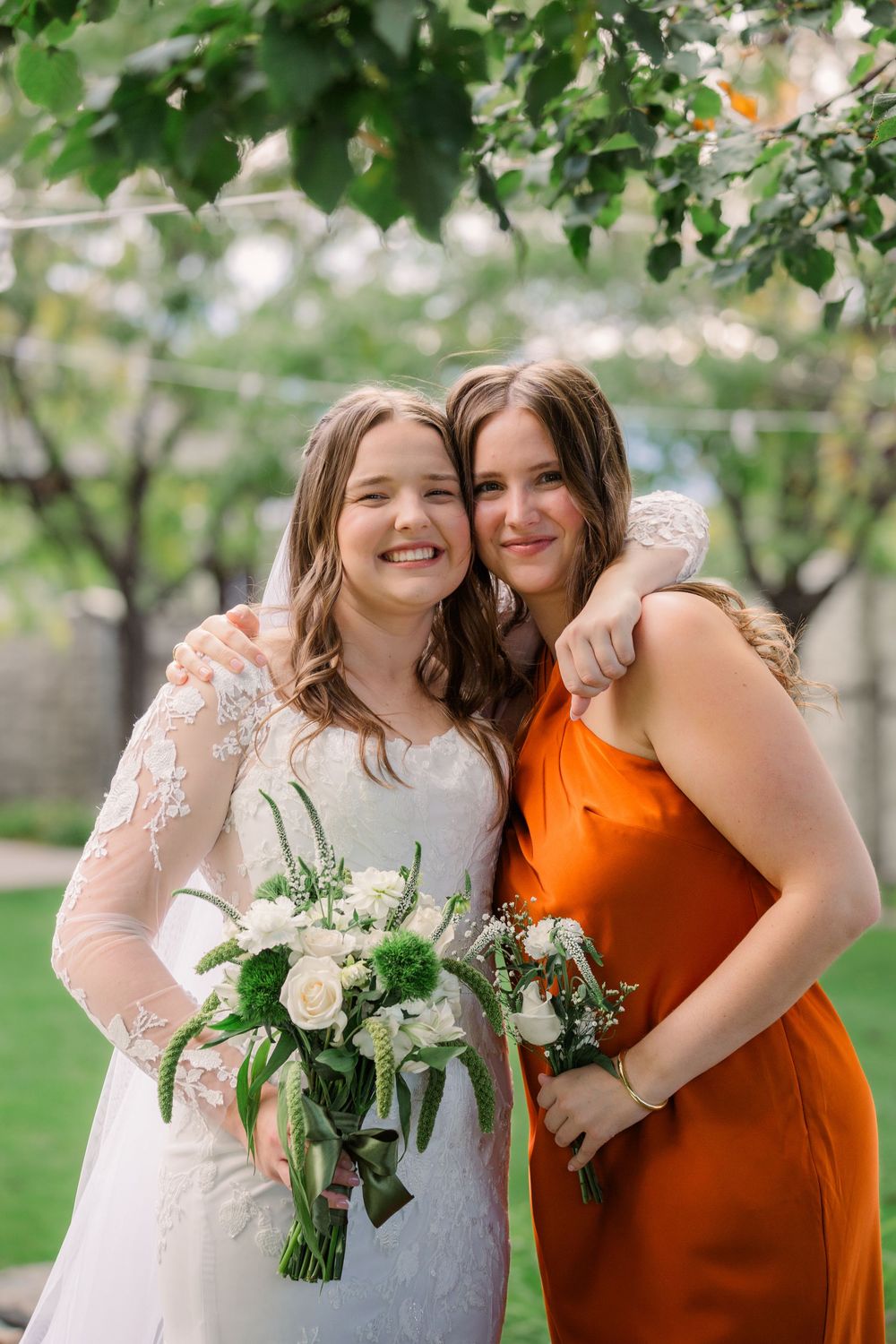A bride in a lace wedding dress and companion in orange dress pose together with white floral bouquet in garden setting.