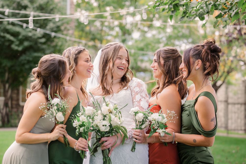 A group of bridesmaids in different colored dresses laugh together while holding bouquets in an outdoor garden setting.