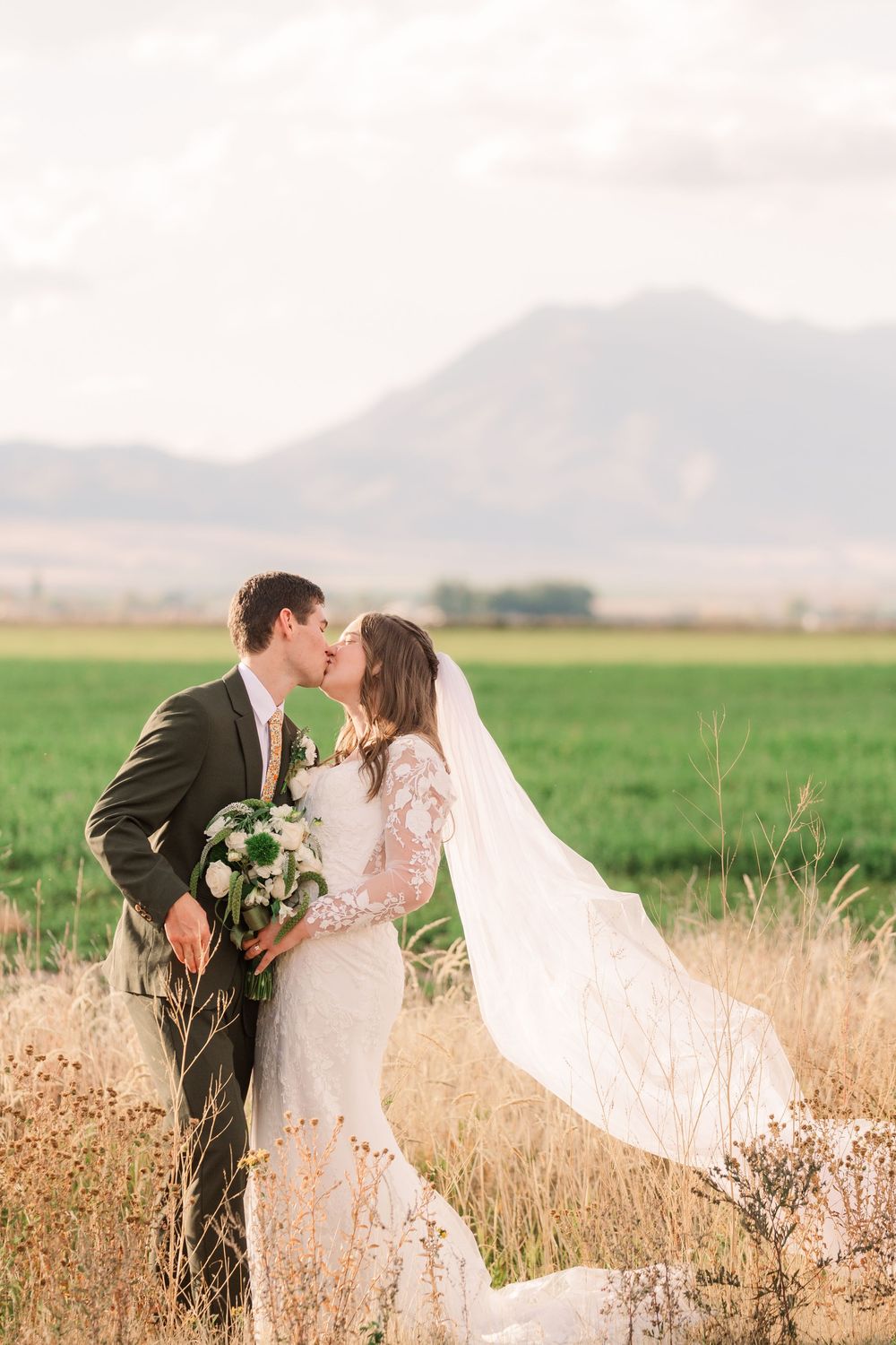 A romantic wedding couple embraces in a golden field with mountains in the background at sunset.