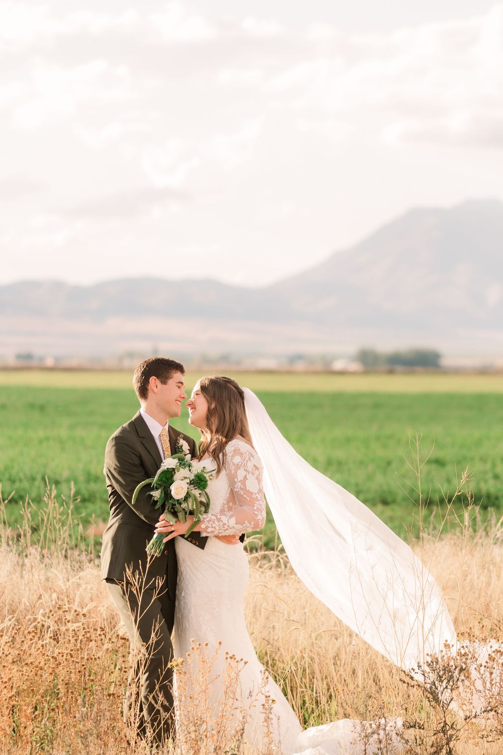 A romantic wedding couple embraces in a golden wheat field as the bride's veil flows dramatically in the wind.