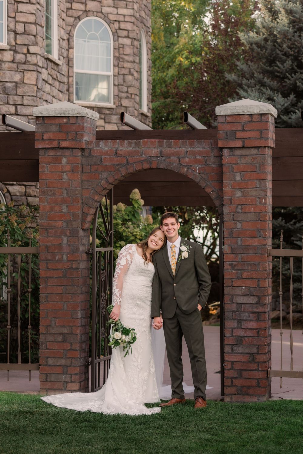 A couple stands under a brick archway entrance at a wedding venue with stone architecture and landscaping.