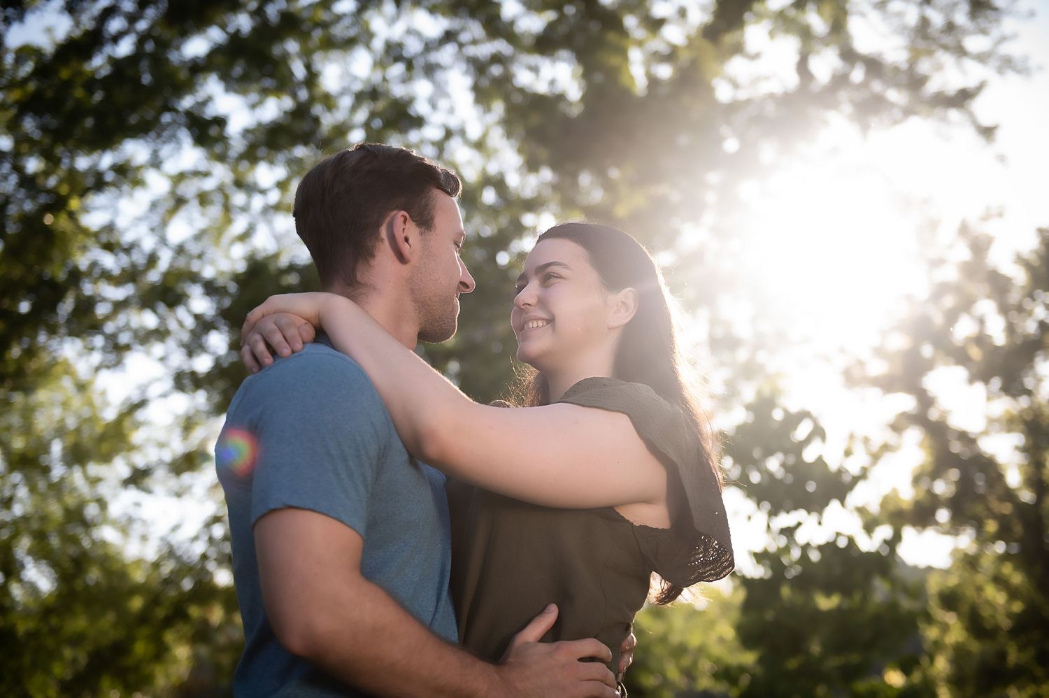 A romantic couple embraces in a sunlit outdoor setting with trees silhouetted against bright sunlight.