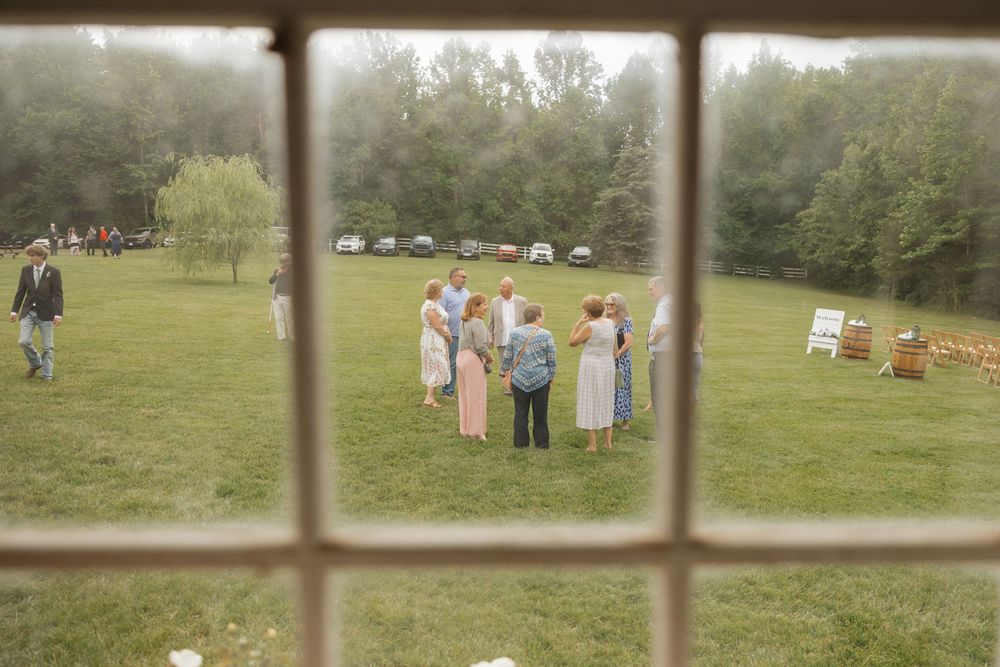 Outdoor wedding ceremony setup viewed through window panes with chairs and guests visible in the distance.