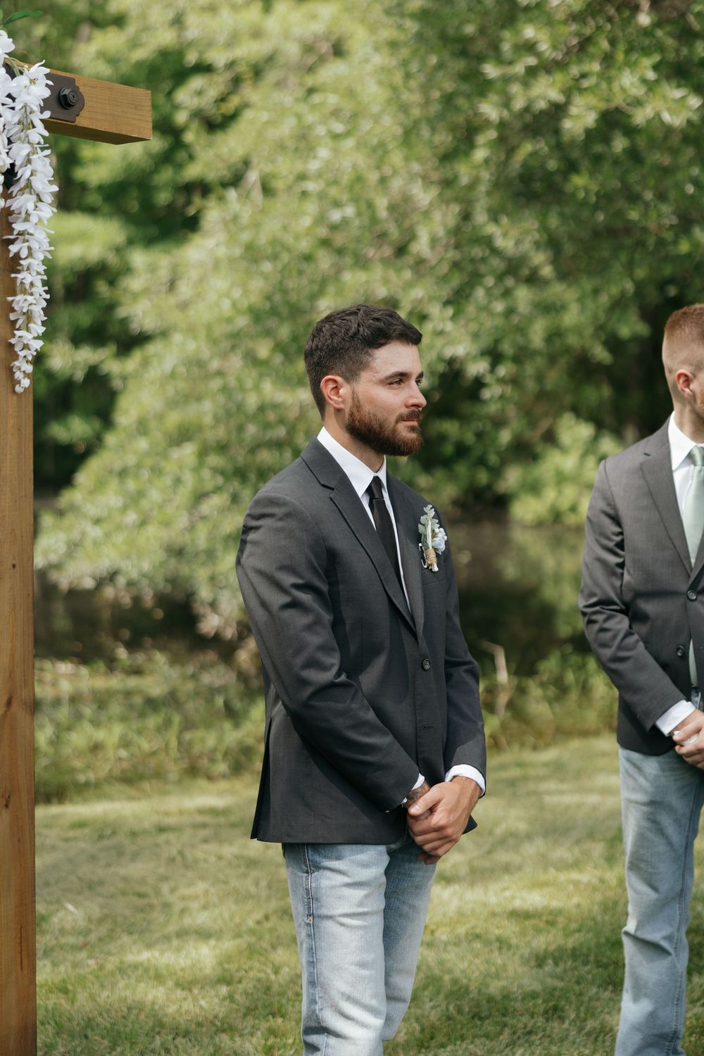 Groomsmen in black suits and light jeans stand together during an outdoor wedding ceremony.